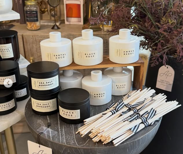 Display of scented candles labeled Santal Coco, Black Rose, Amber Berry, Sea Salt Bergamot, and Black Rose, along with white and black striped reed diffusers or aroma sticks on a dark round table.
