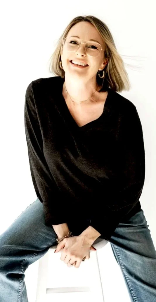 A young woman with shoulder-length blond hair, wearing glasses, a black top, and jeans, smiling and sitting on a white stool against a white background.