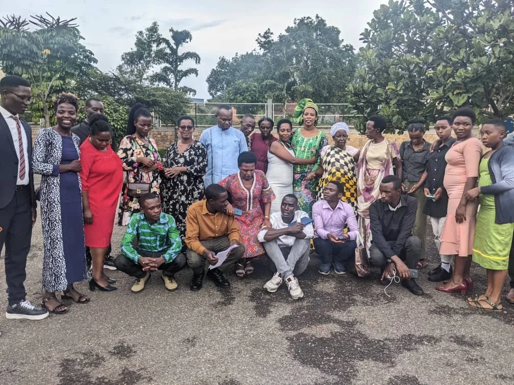 A group of people, mostly women, standing and sitting outdoors on a wet road, some holding papers and phones, with trees and fencing in the background.