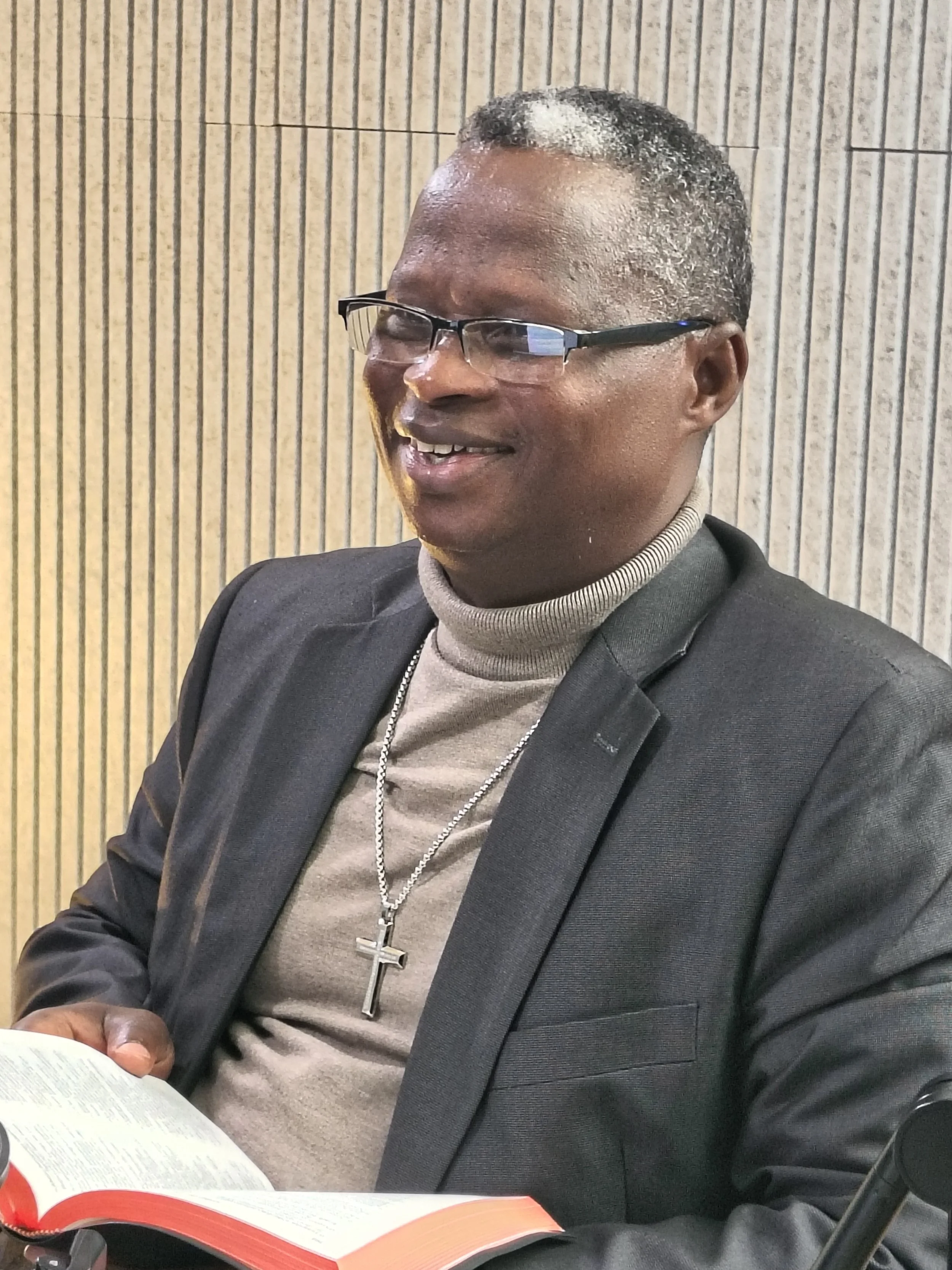 A man with black hair and glasses sitting at a table, reading a book. He wears a gray turtleneck and a dark blazer, with a silver necklace and cross pendant. The background has vertical striped wallpaper.