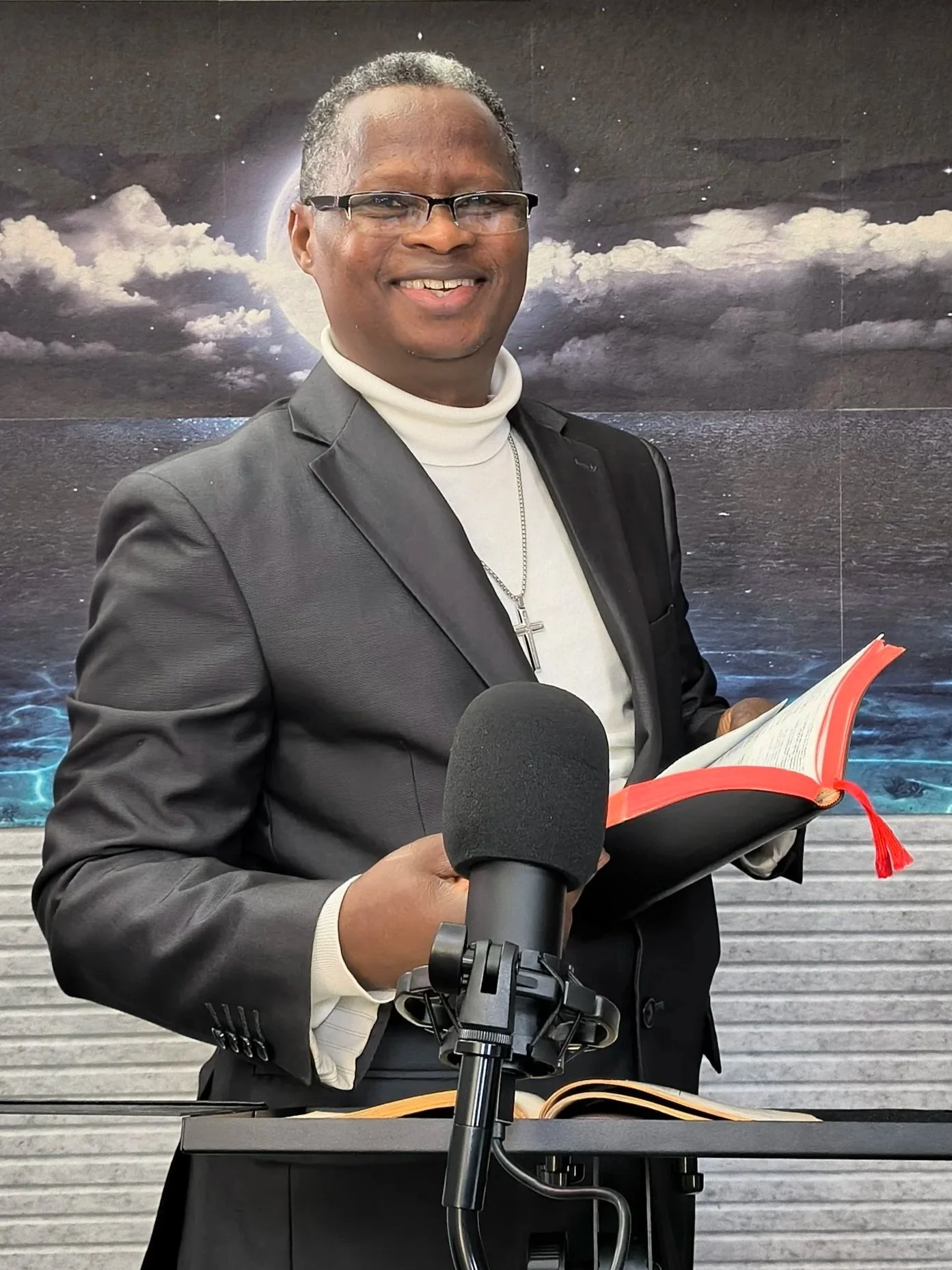 A man in a suit holding a microphone and reading from a red-covered book, standing in front of a scenic mountain and water background.