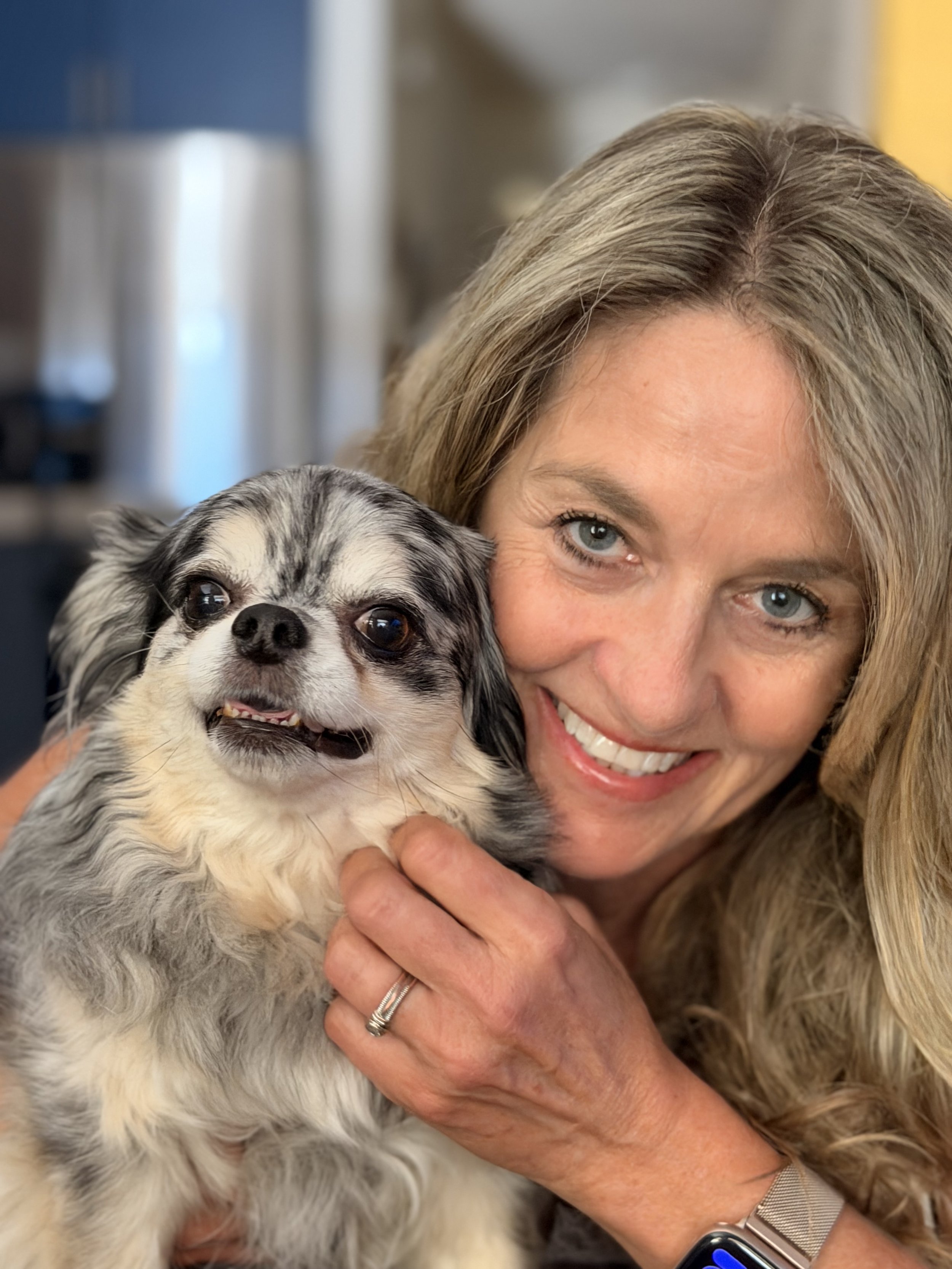 A woman with blonde hair and blue eyes holding a small, fluffy, black and white dog close to her face, smiling.
