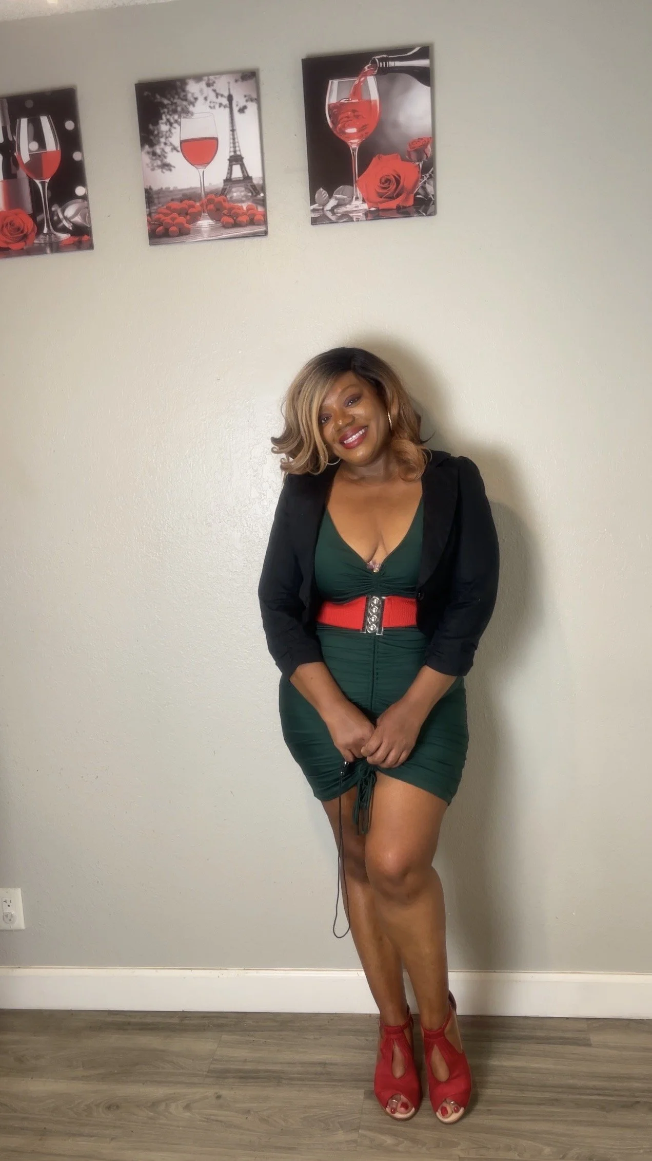 A woman in a green dress with a red belt and red high-heeled shoes standing in front of a plain wall with three framed pictures of wine, roses, and Paris.