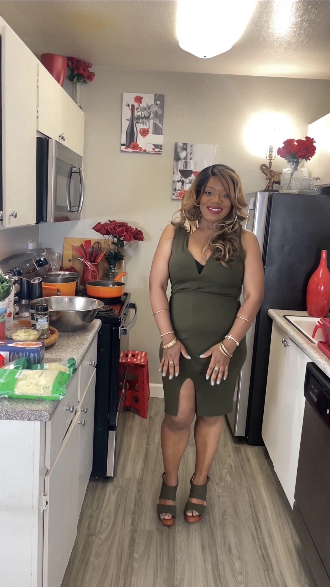 A woman in a green dress standing in a kitchen, smiling at the camera. The kitchen has white cabinets, gray countertops, and various cooking utensils and decorations, including red flowers and artwork on the wall.