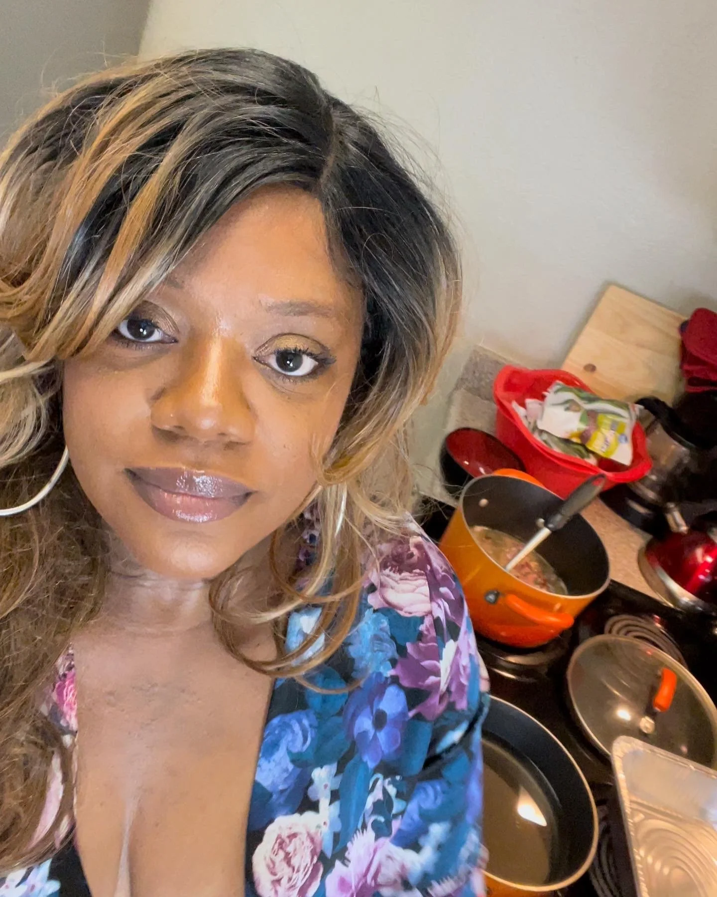 A woman with light brown and black hair styled in loose waves, wearing makeup with gold eyeshadow, standing in a kitchen. In the background, there are pots, pans, a red bowl, and a counter with various items.