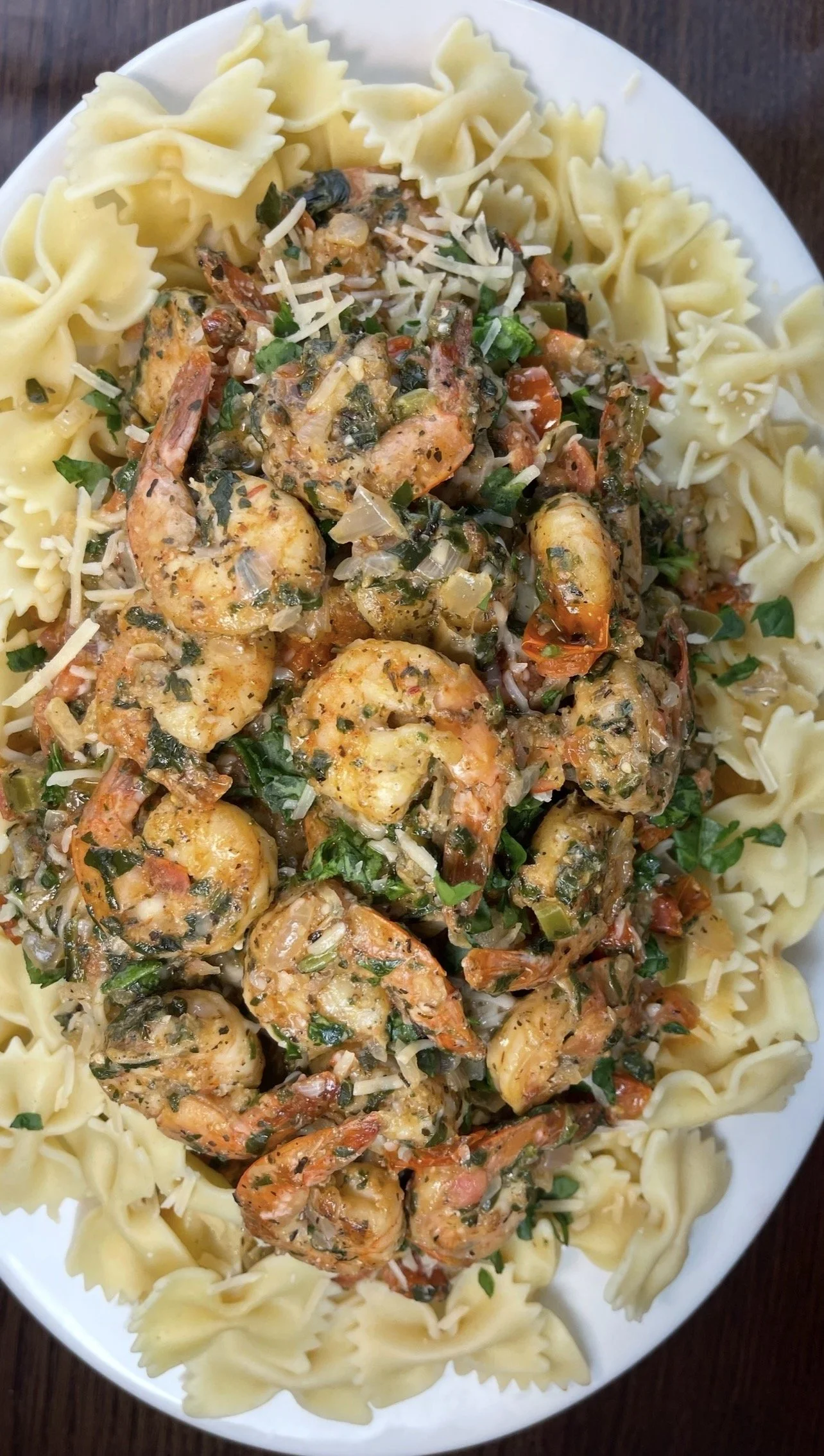 A plate of cooked bowtie pasta topped with shrimp and a herb sauce, garnished with shredded cheese and chopped greens.