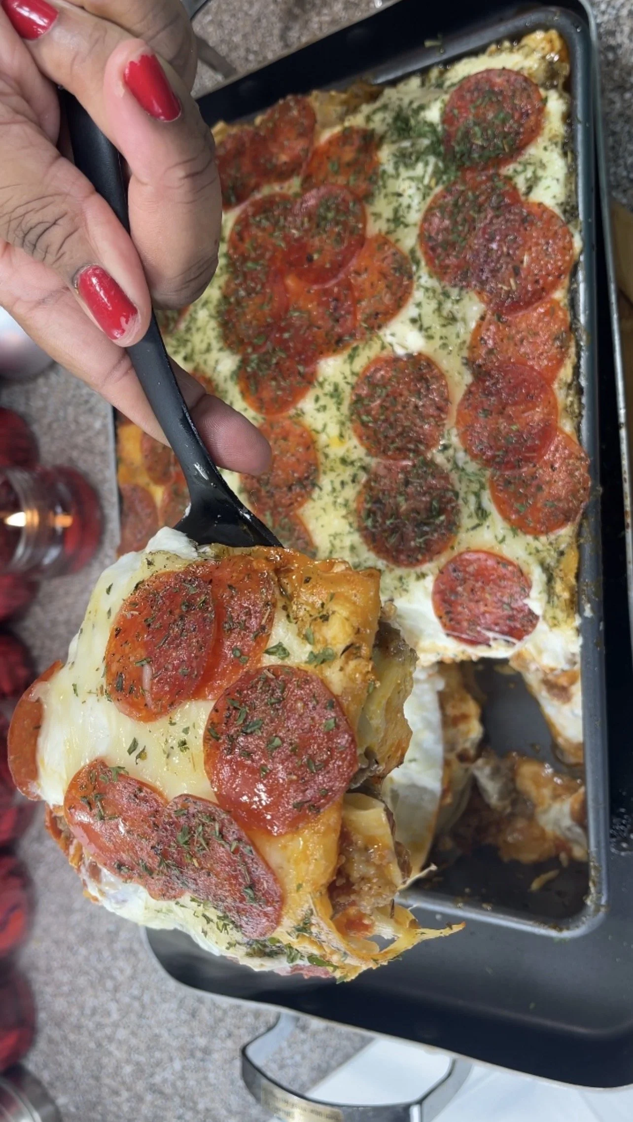 Close-up of a slice of pepperoni pizza with melted cheese, herbs, and toppings being lifted out of a baking pan, with a hand holding the slice.