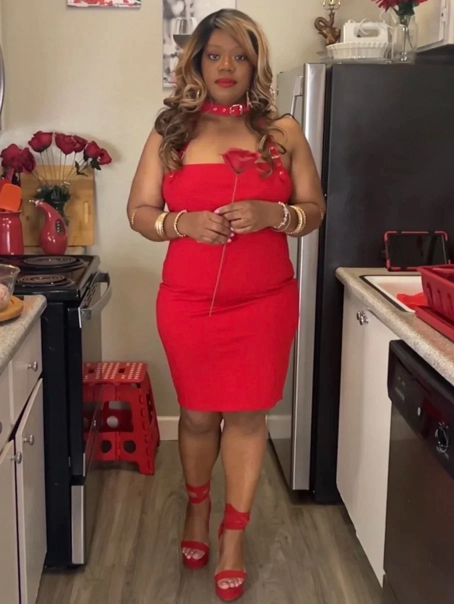 A woman standing in a kitchen wearing a red dress and high heels, holding a red rose, with red and gold jewelry and curly hair.