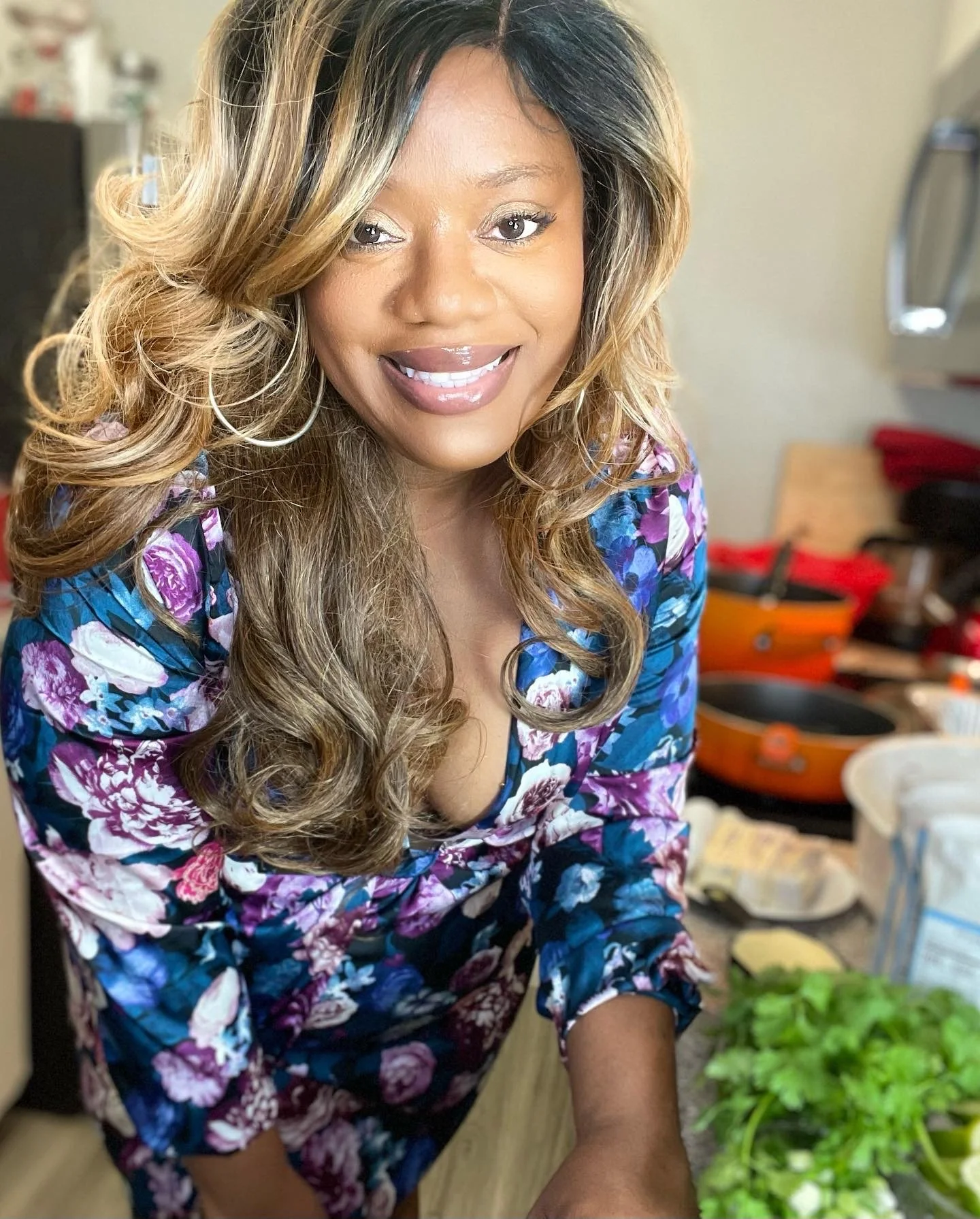 Smiling woman with long wavy hair, wearing a floral blouse, standing in a kitchen with fresh vegetables and cooking supplies visible.