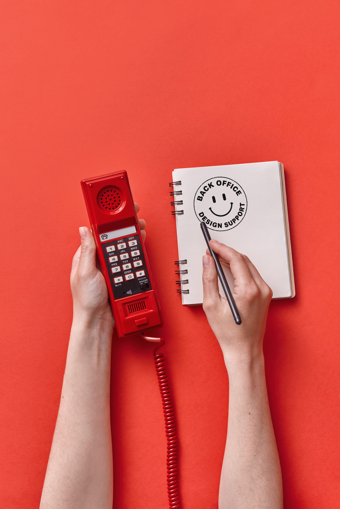 A person holding a red emergency phone in one hand and writing on a notepad with the other hand on a red background, with the notepad displaying a smiley face and the words 'Back Office Design Support'.