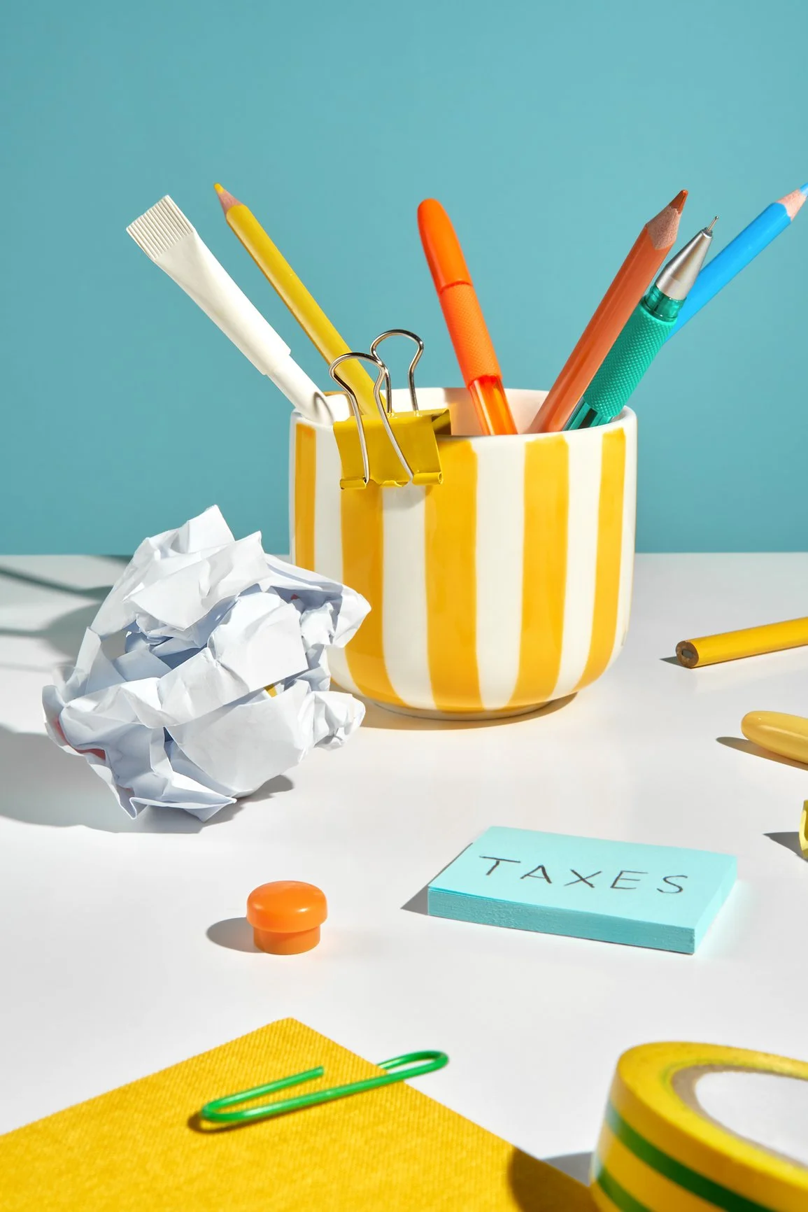 Desk with crumpled paper, sticky notes labeled 'TAXES', and various office supplies like pens in a yellow and white striped cup against a blue background.