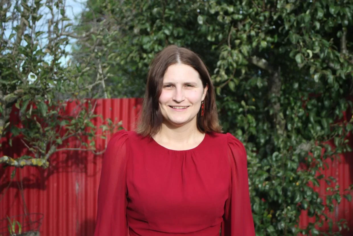 A woman with shoulder-length brown hair wearing a red dress standing outdoors in front of a red fence and green foliage.