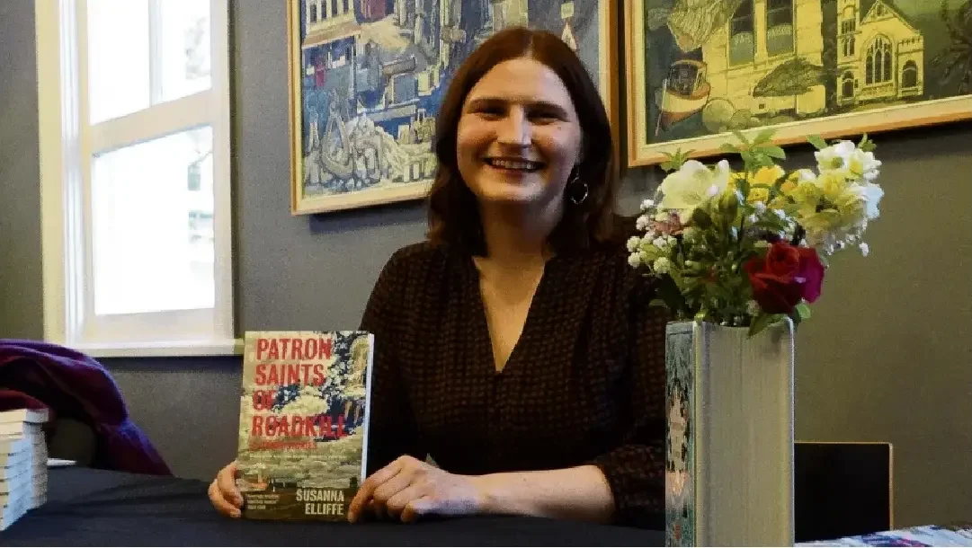 Woman with shoulder-length dark hair smiling, sitting at a table with a book titled 'Patron Saints of Rebelling,' and a vase with white and red flowers, indoor setting with artworks on the wall.