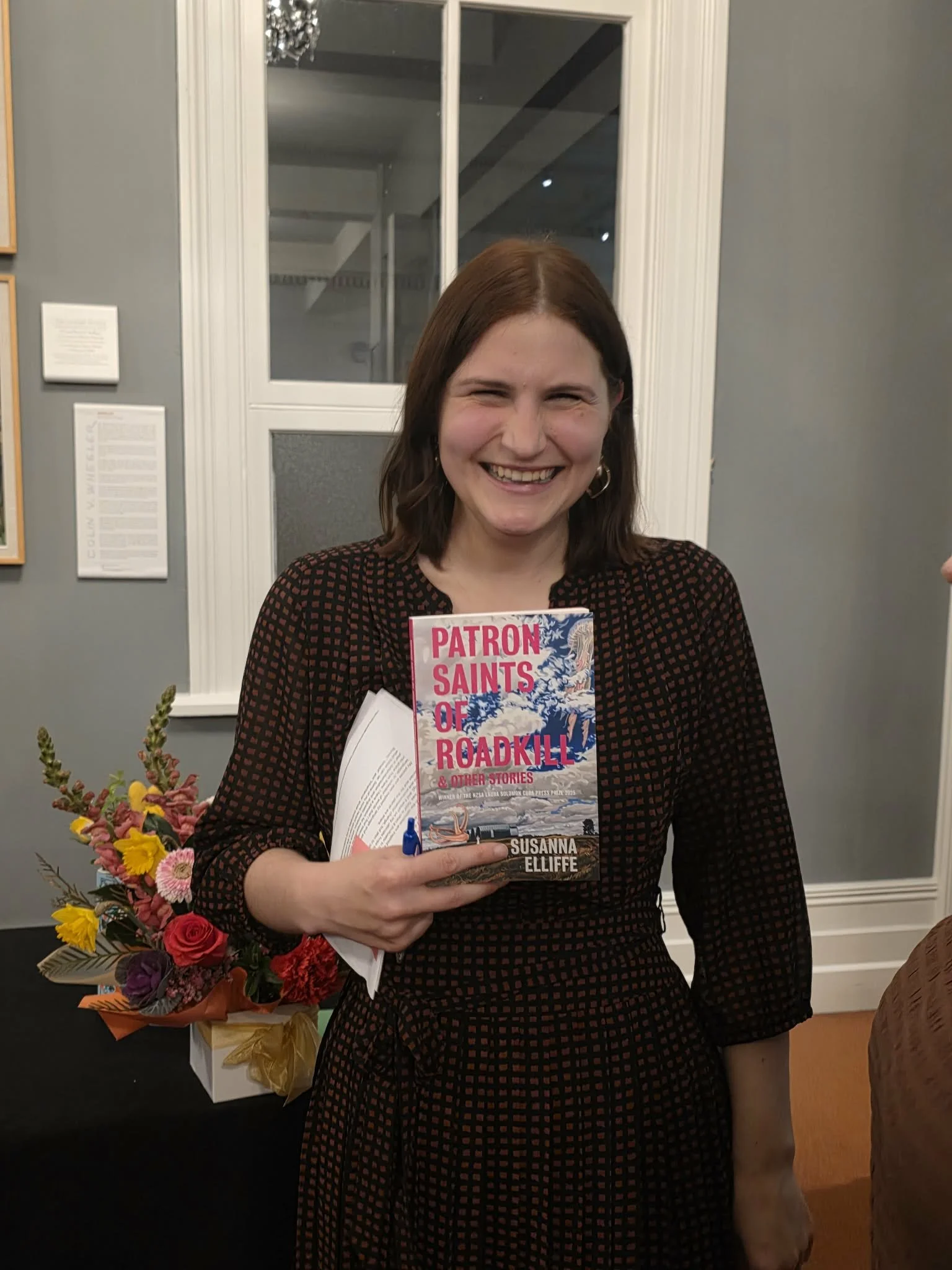 A woman with shoulder-length brown hair, wearing a black dress with red checkered pattern, is smiling and holding a book titled 'Patron Saints of Roadkill & Other Stories' by Susanna Elliffe. She is standing indoors with a window behind her, beside a bouquet of colorful flowers on a table.