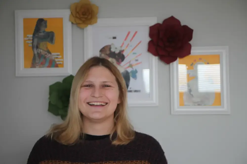 Smiling woman with blonde hair standing in front of a wall decorated with colorful framed artwork and large artificial paper flowers.