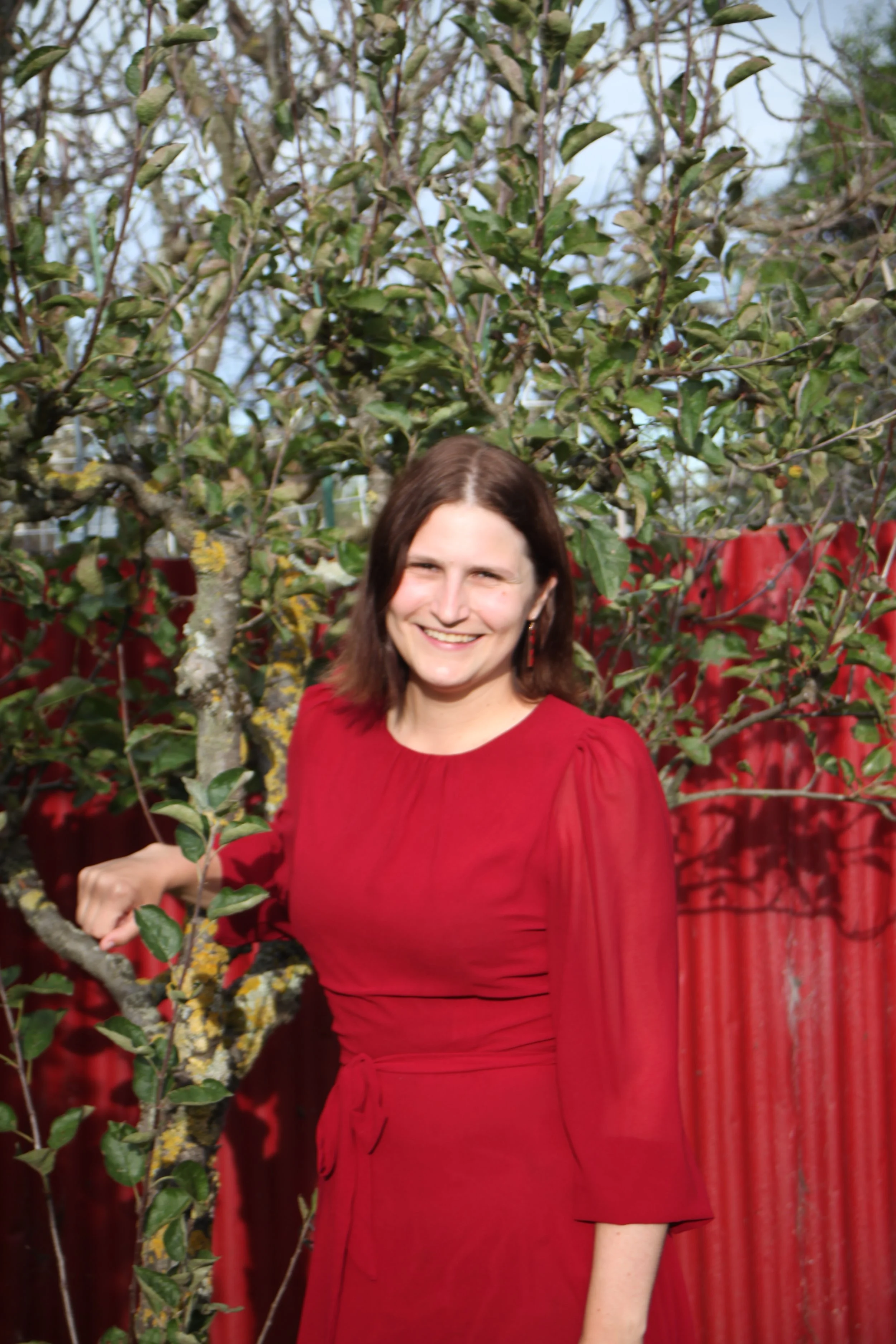 A woman in a red dress standing outdoors in front of a red fence and leafy trees, smiling.