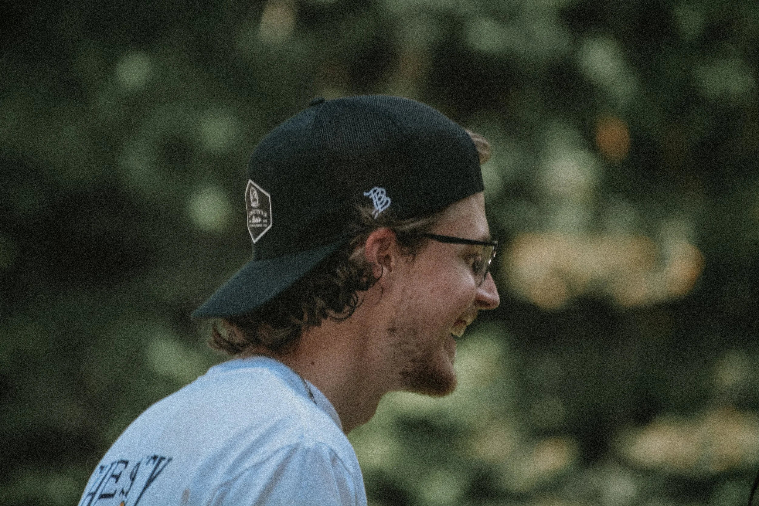 A young person with curly hair, wearing a black cap and glasses, smiling and looking to the side outdoors.