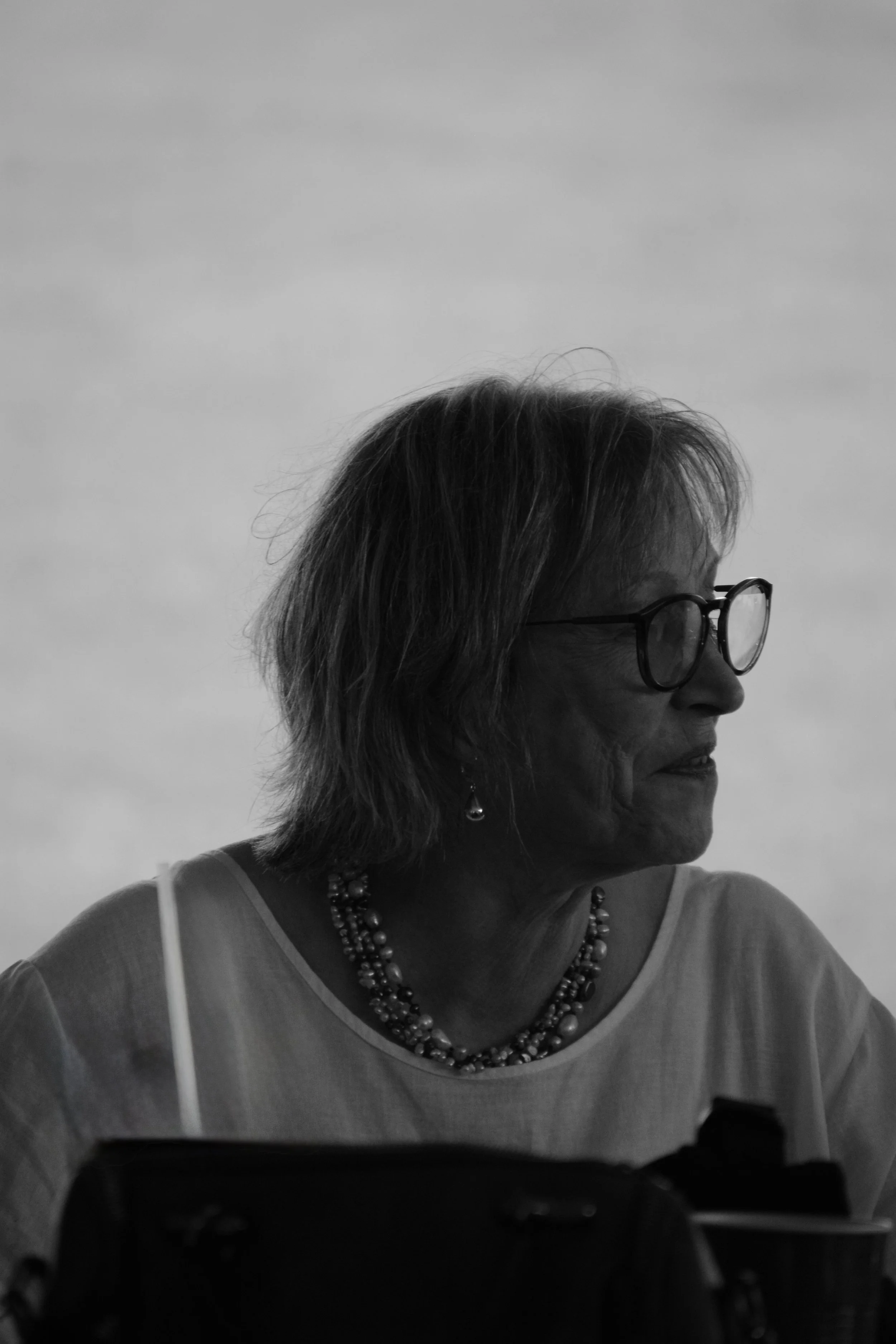 Black and white photo of a woman with glasses and short hair, looking to her left, wearing a necklace and a top with thin straps, sitting indoors with a cloudy sky background.