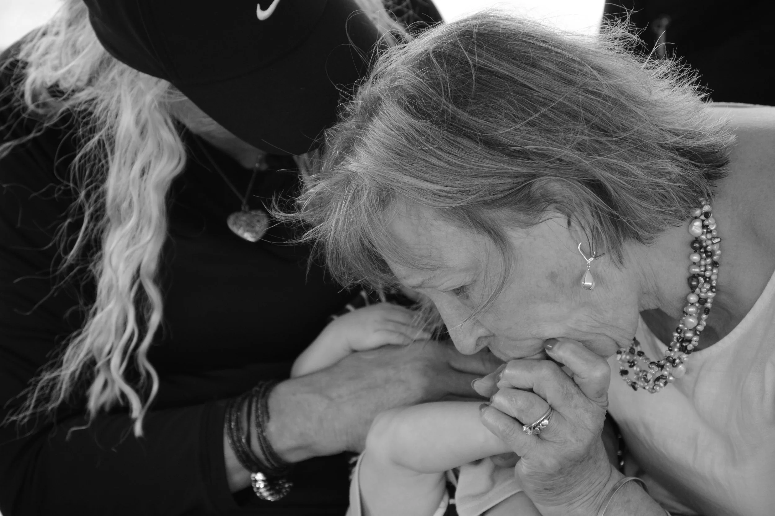 An elderly woman with short hair, wearing earrings and multiple necklaces, is being comforted, possibly crying, with her head bowed and hands covering her face as a person in dark clothing and a hat consoles her.