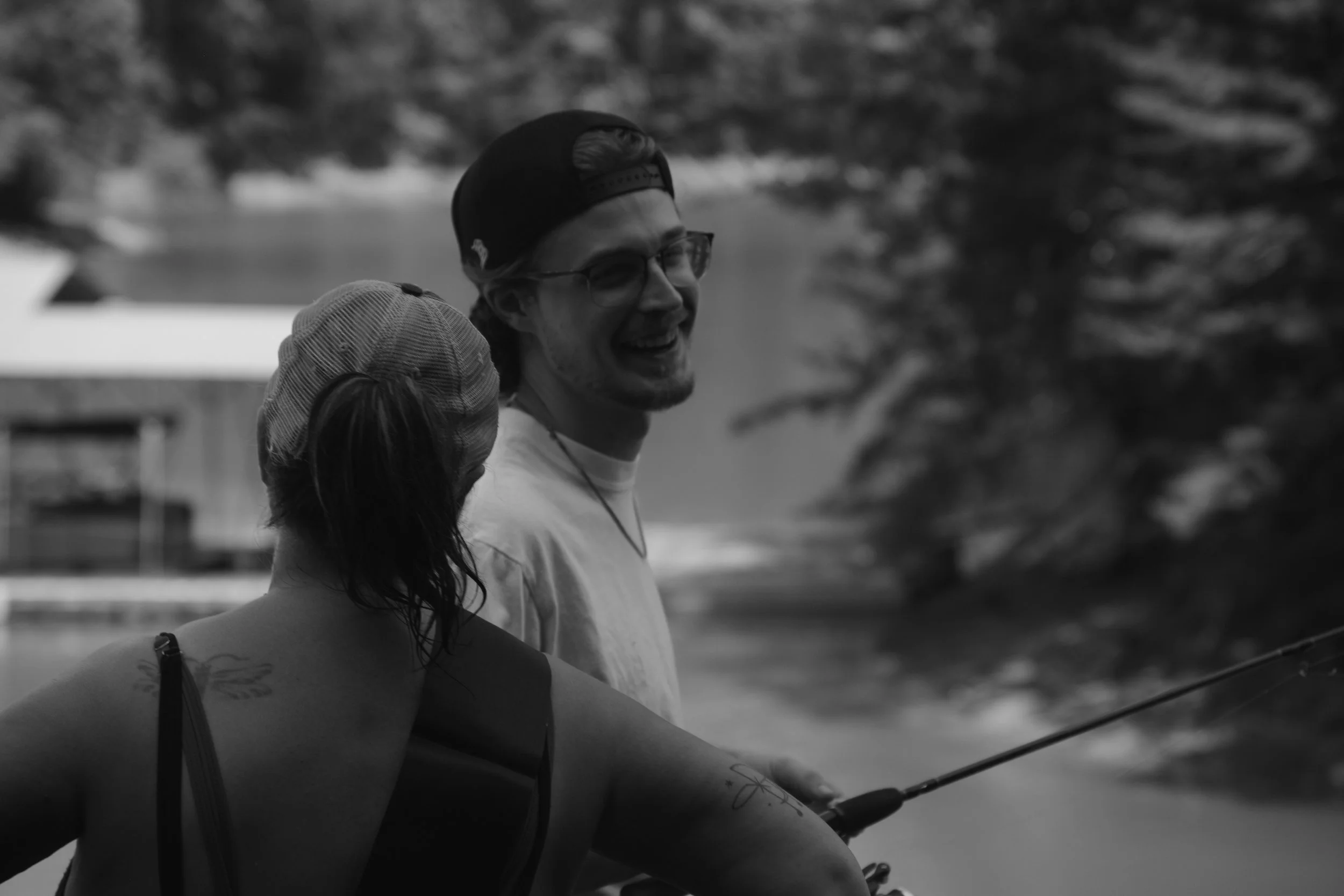 A person smiling while sharing a moment with someone else, outdoor setting with blurry trees and structures in background, black and white photo.