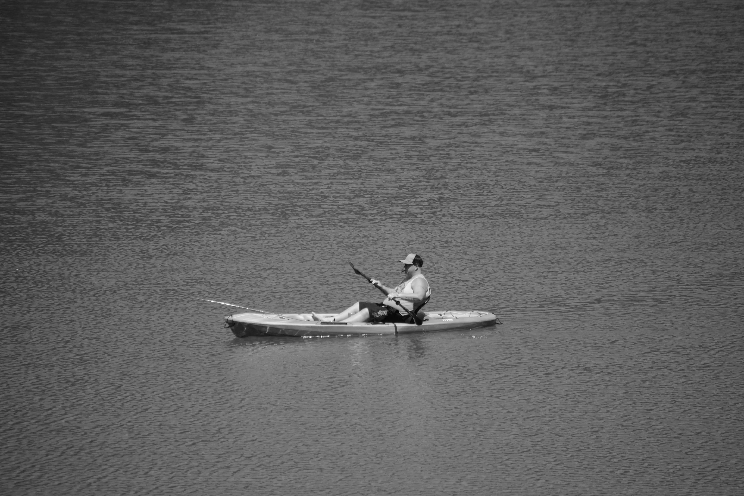 A person sitting in a kayak on a large, calm body of water, wearing sunglasses and a white cap.