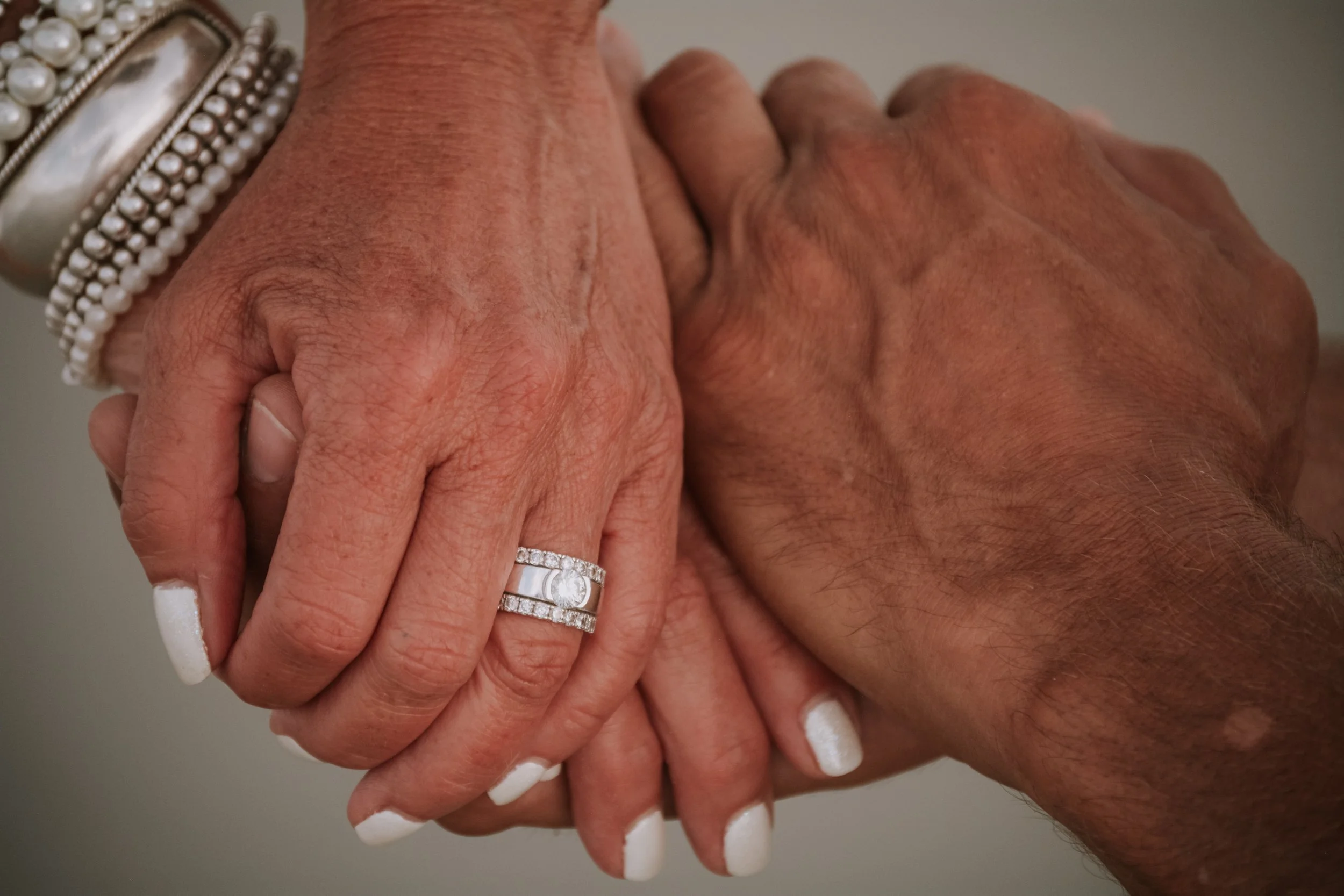 Two people holding hands, one wearing a pearl bracelet and diamond rings, the other with a dark skin tone and a silver bracelet.