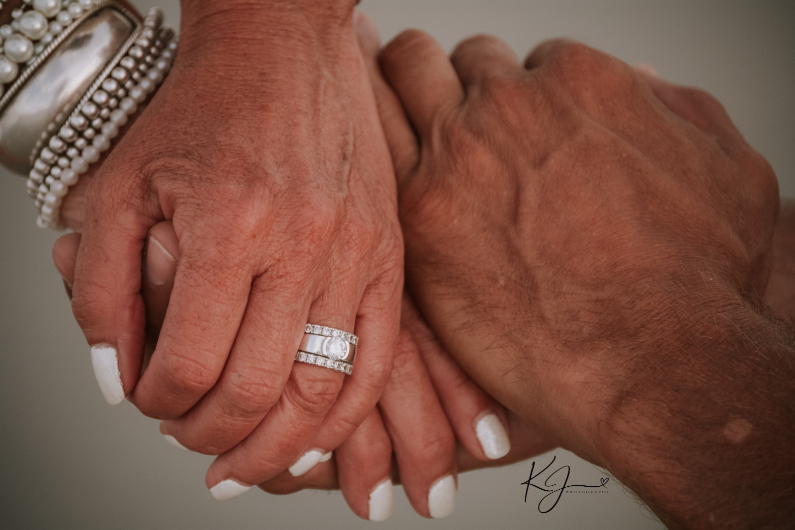 Close-up of two hands clasped, one with wedding ring and white painted nails, the other with a bracelet, suggesting an intimate or romantic moment.