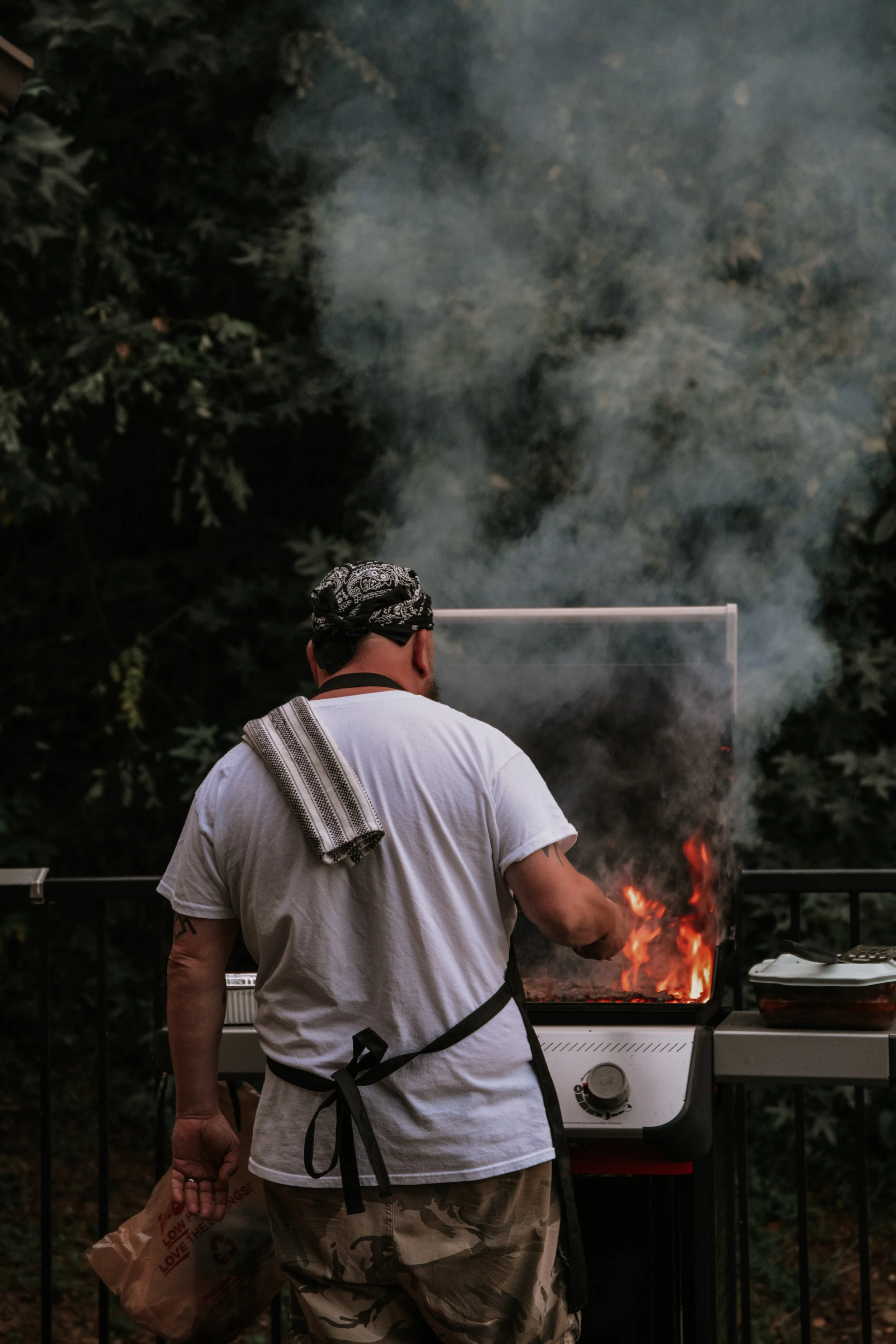 A person grilling outdoors at night, wearing a bandana, white T-shirt, and camouflage shorts, holding a paper bag, with smoke and flames from the grill.