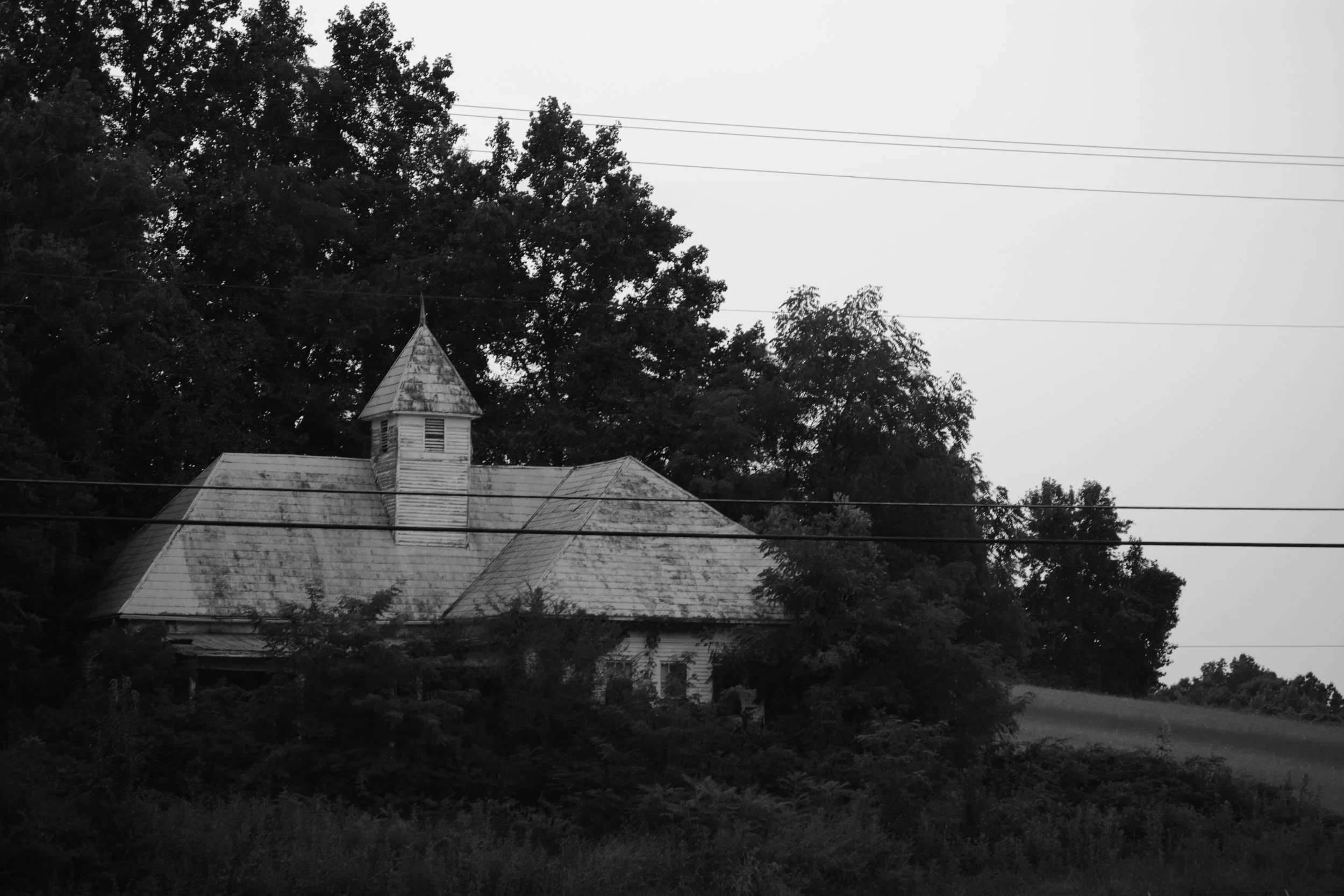 A black and white photo of a house with a steep roof and a small steeple, surrounded by trees, with power lines crossing the sky above.