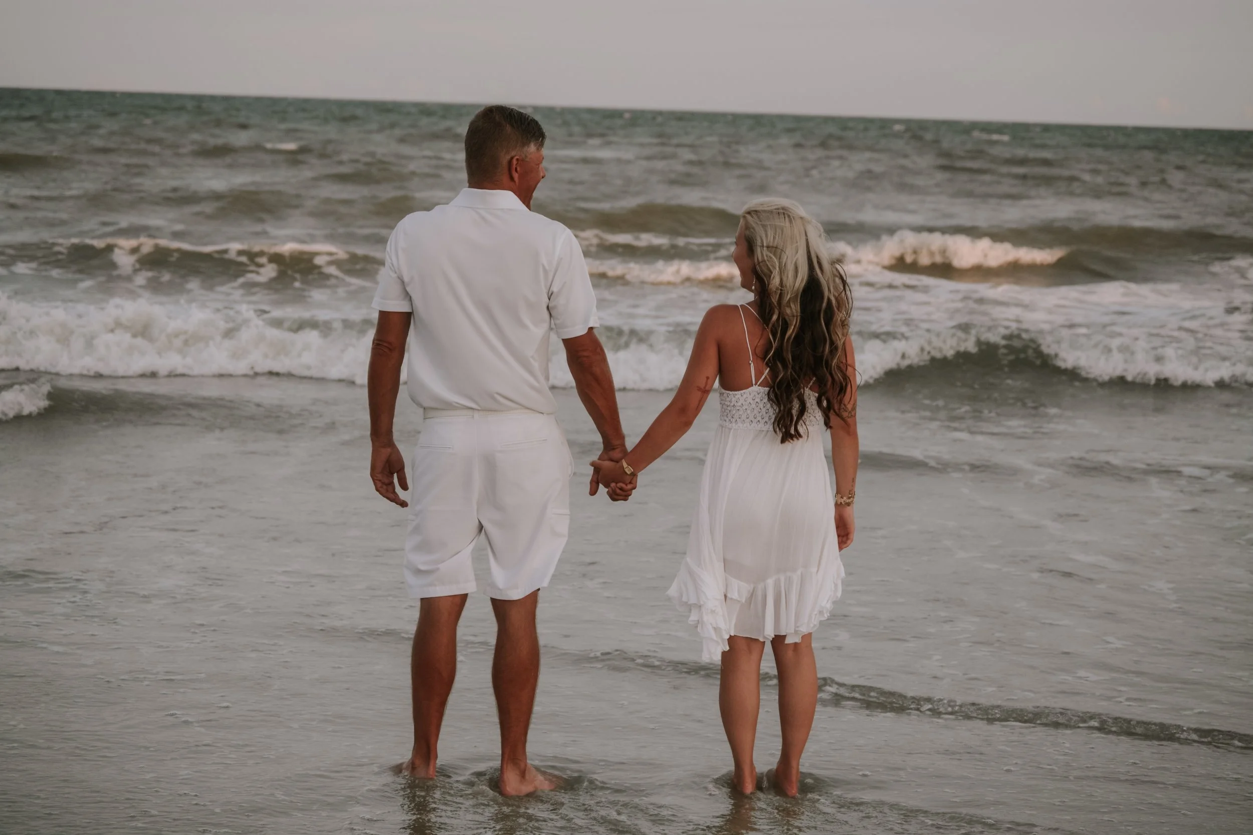 An elderly man and a young woman holding hands and walking on the beach near the ocean, dressed in white summer clothing.