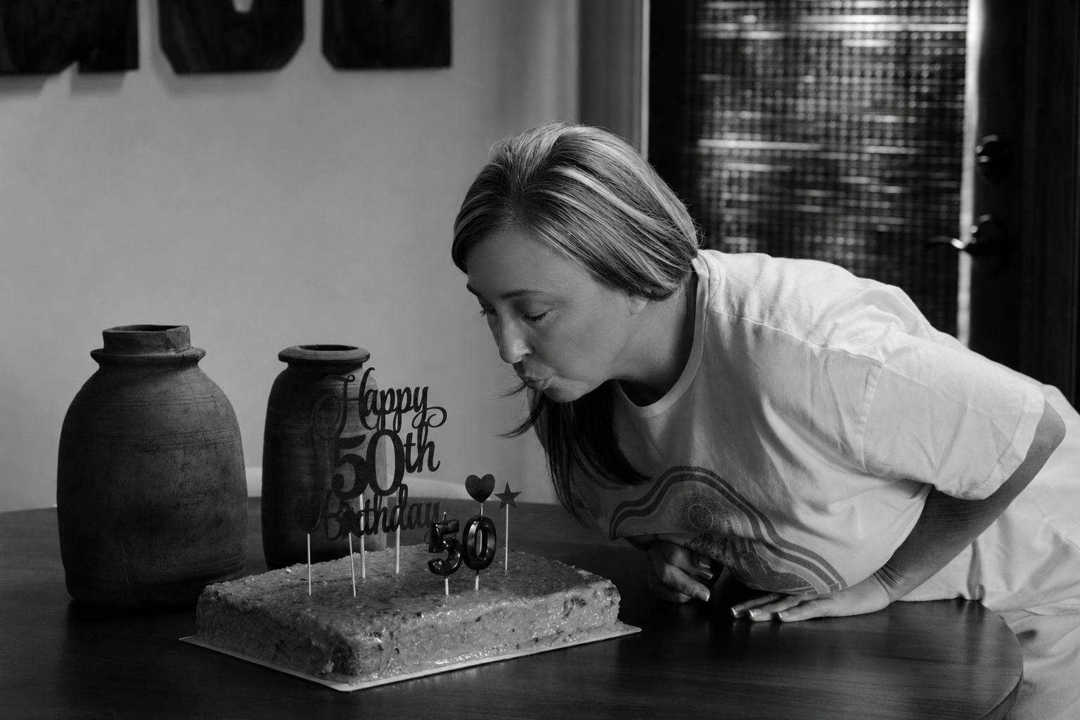 A woman blowing out candles on a birthday cake decorated with the number 50 and a «Happy 50th Birthday» topper, with two vases on the table, celebrating her 50th birthday.