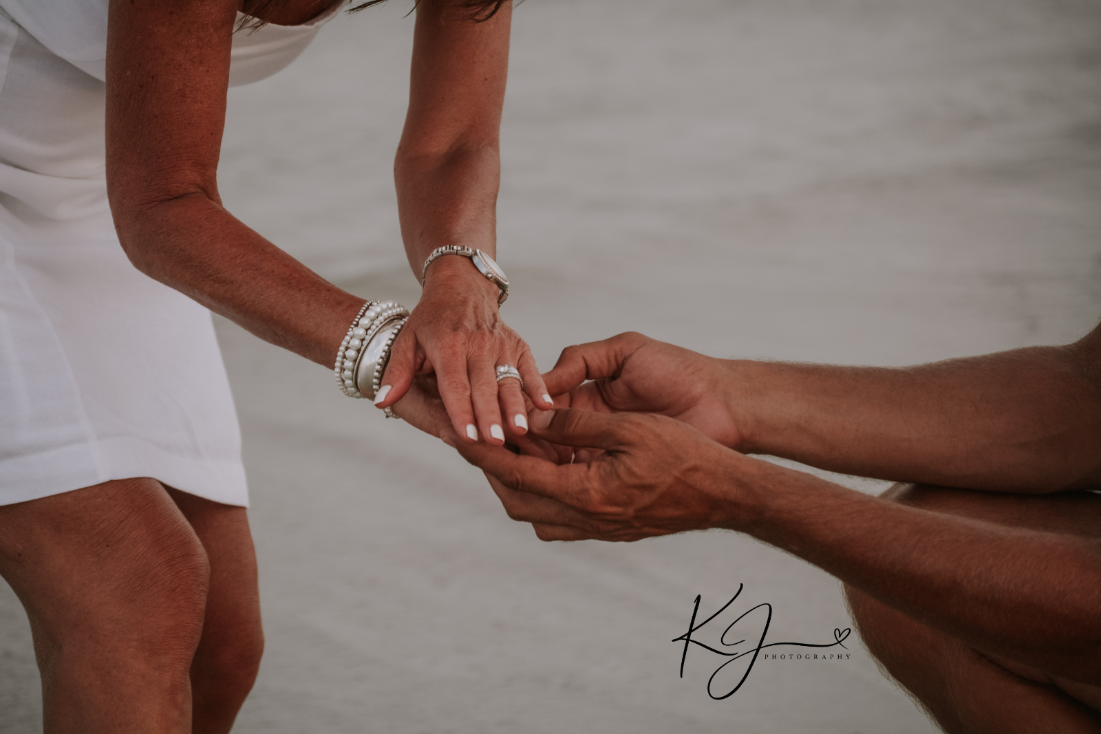 A woman is holding hands with a man who is kneeling on the ground with a beach in the background, suggesting a proposal or romantic moment.