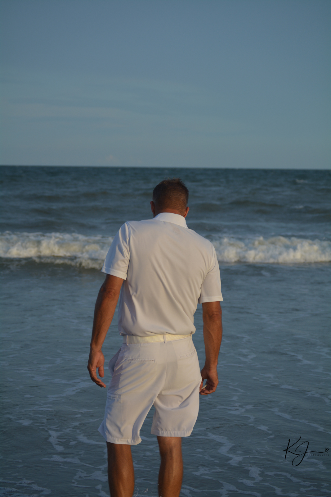 Man in a white shirt and shorts standing in the ocean near the shore with waves and a blue sky in the background.