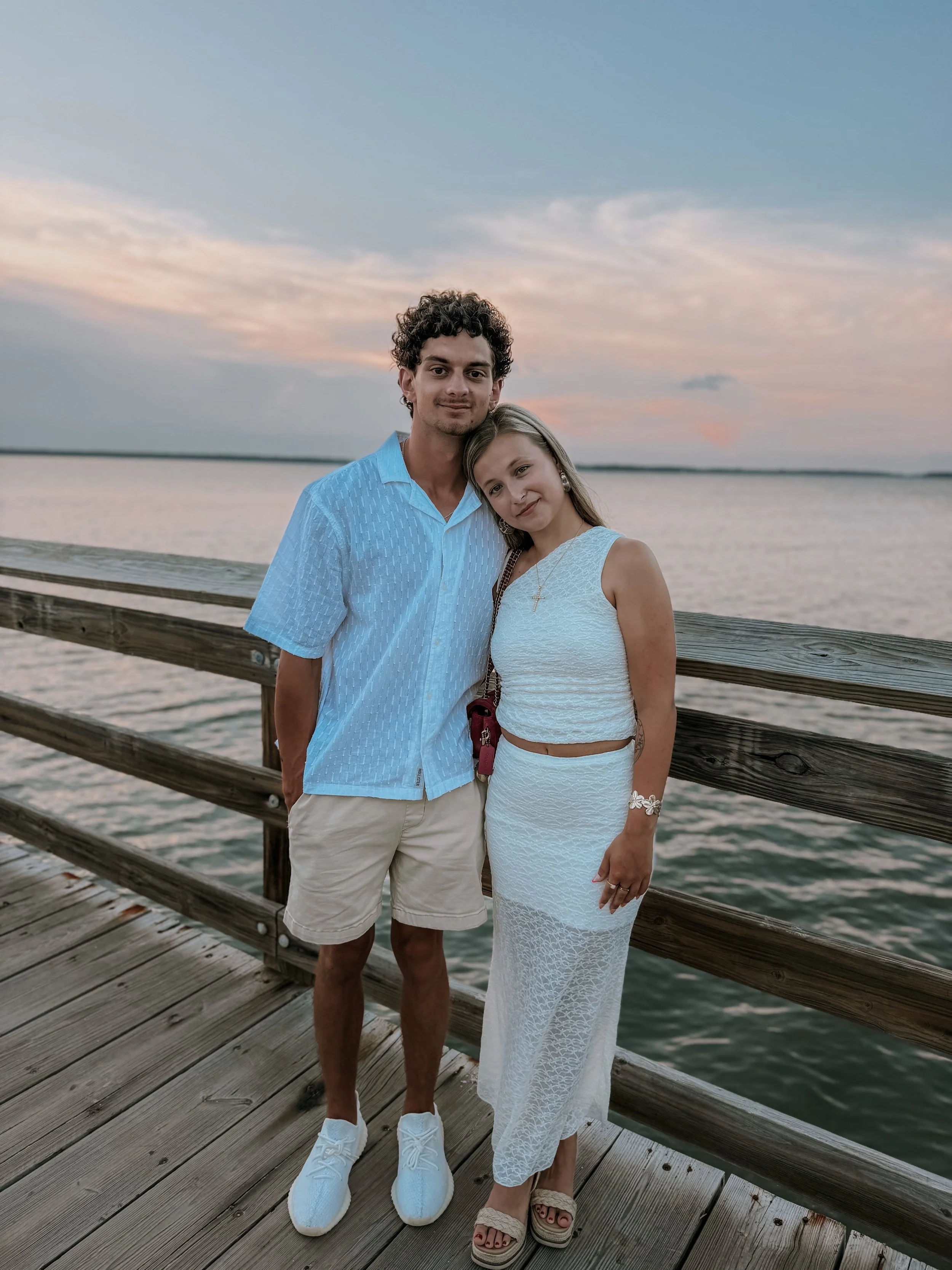 A young man and woman stand closely together on a wooden pier by water, with a sky at sunset in the background. The man is wearing a light blue short-sleeved button-up shirt, beige shorts, and white sneakers. The woman is dressed in a white sleeveless textured dress and tan wedge sandals, with her head resting on the man's shoulder.
