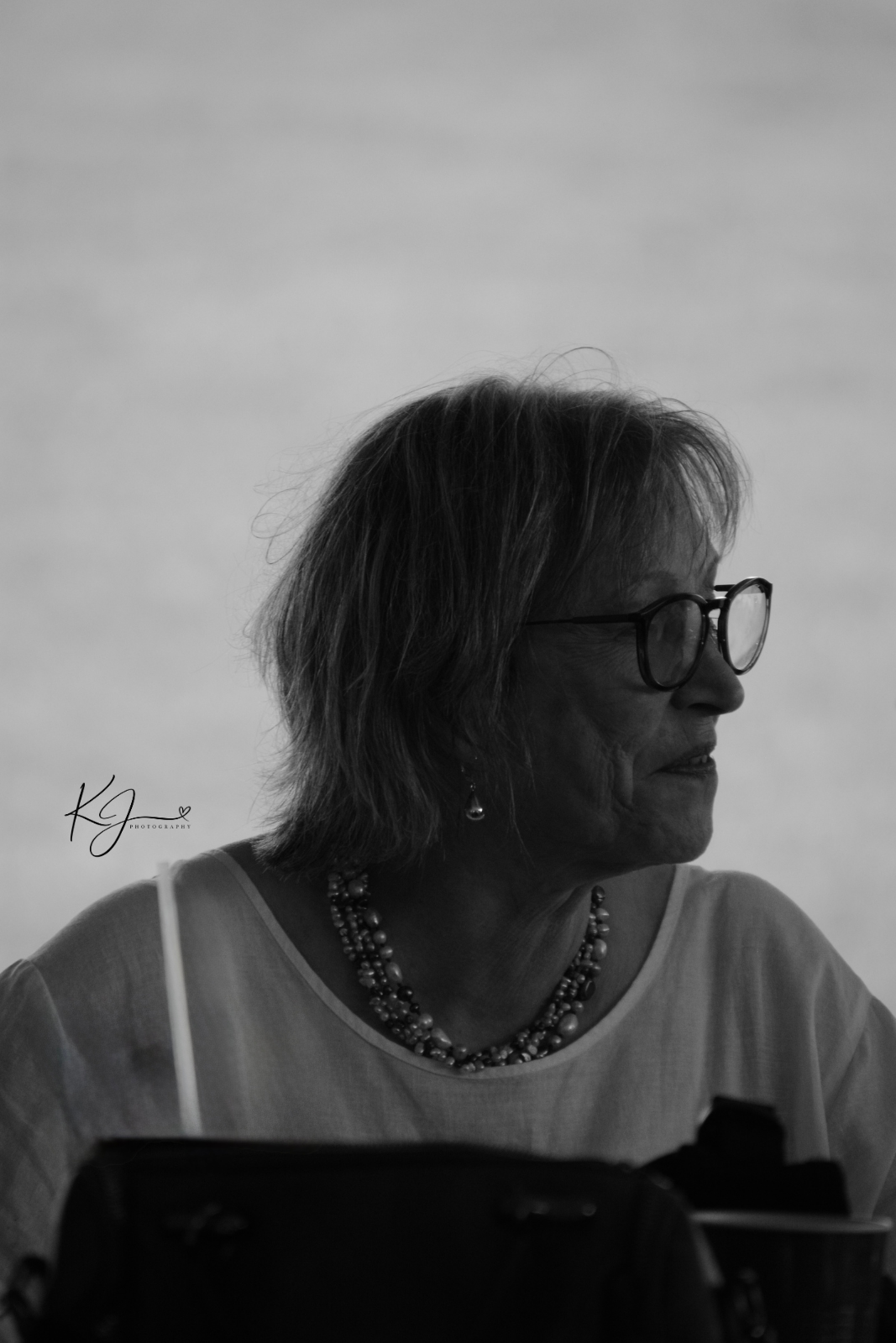 Side profile of an elderly woman with glasses, wearing earrings and a beaded necklace, looking to her right.