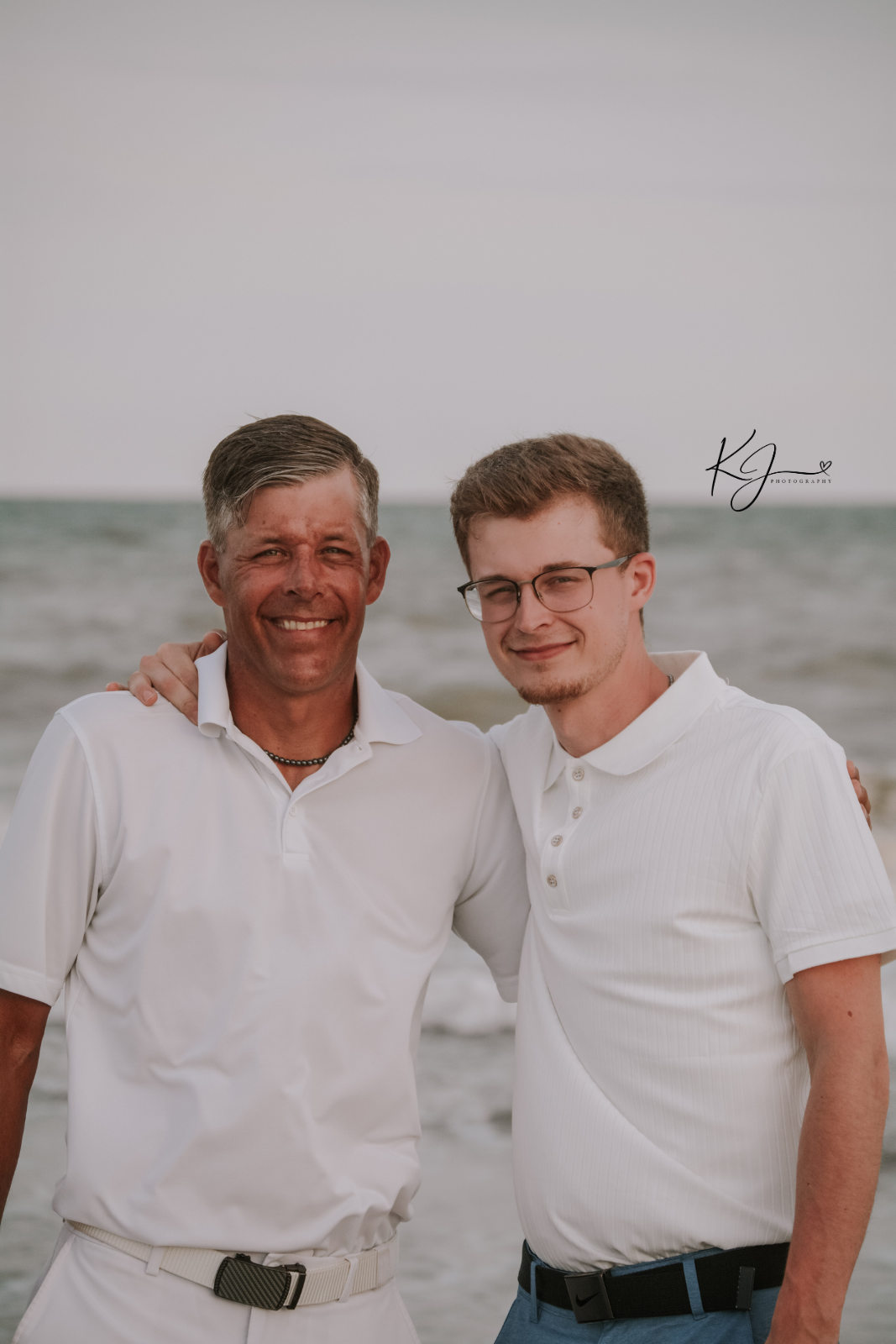 Two men standing on a beach with ocean waves in the background, smiling and embracing each other.