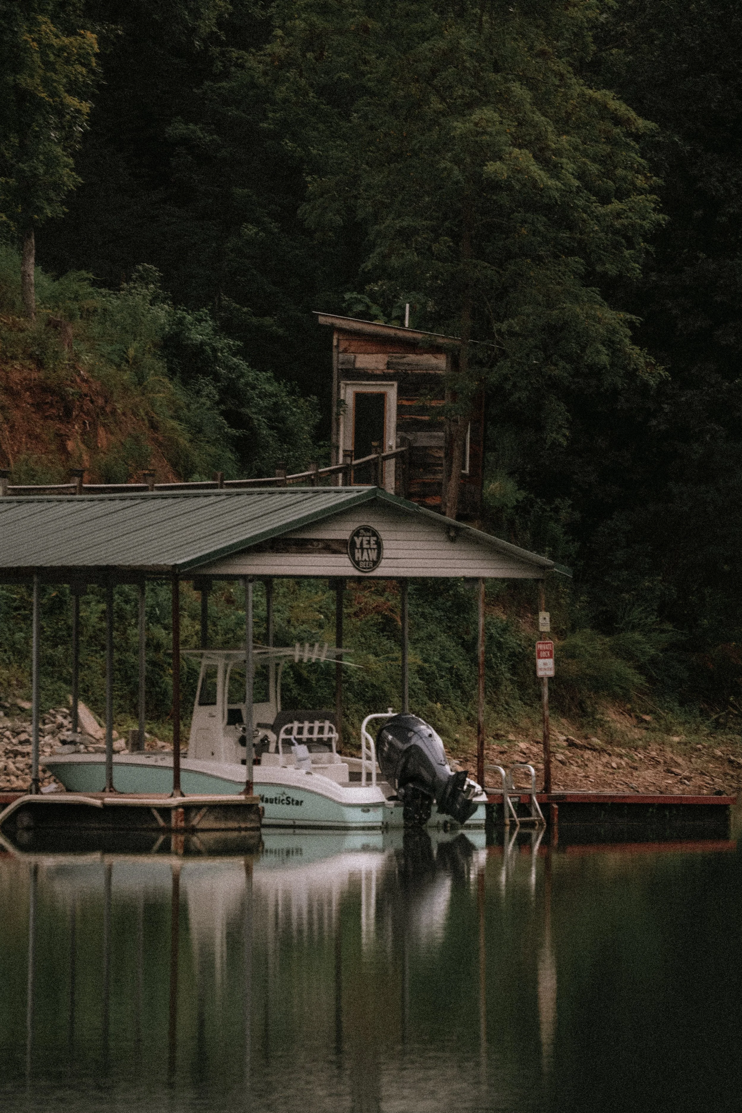 A docked boat on a small waterbody with a covered seating area and some chairs, with a wooded hillside and buildings in the background.