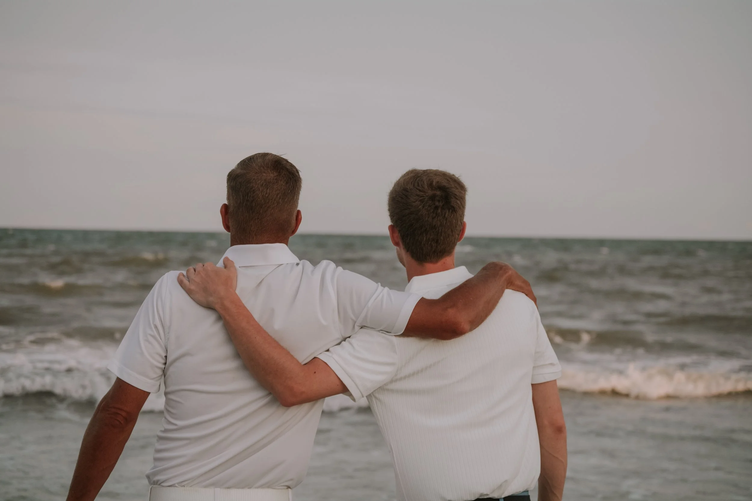 Two men with short hair wearing white shirts and embracing while standing on the beach, looking out at the ocean.