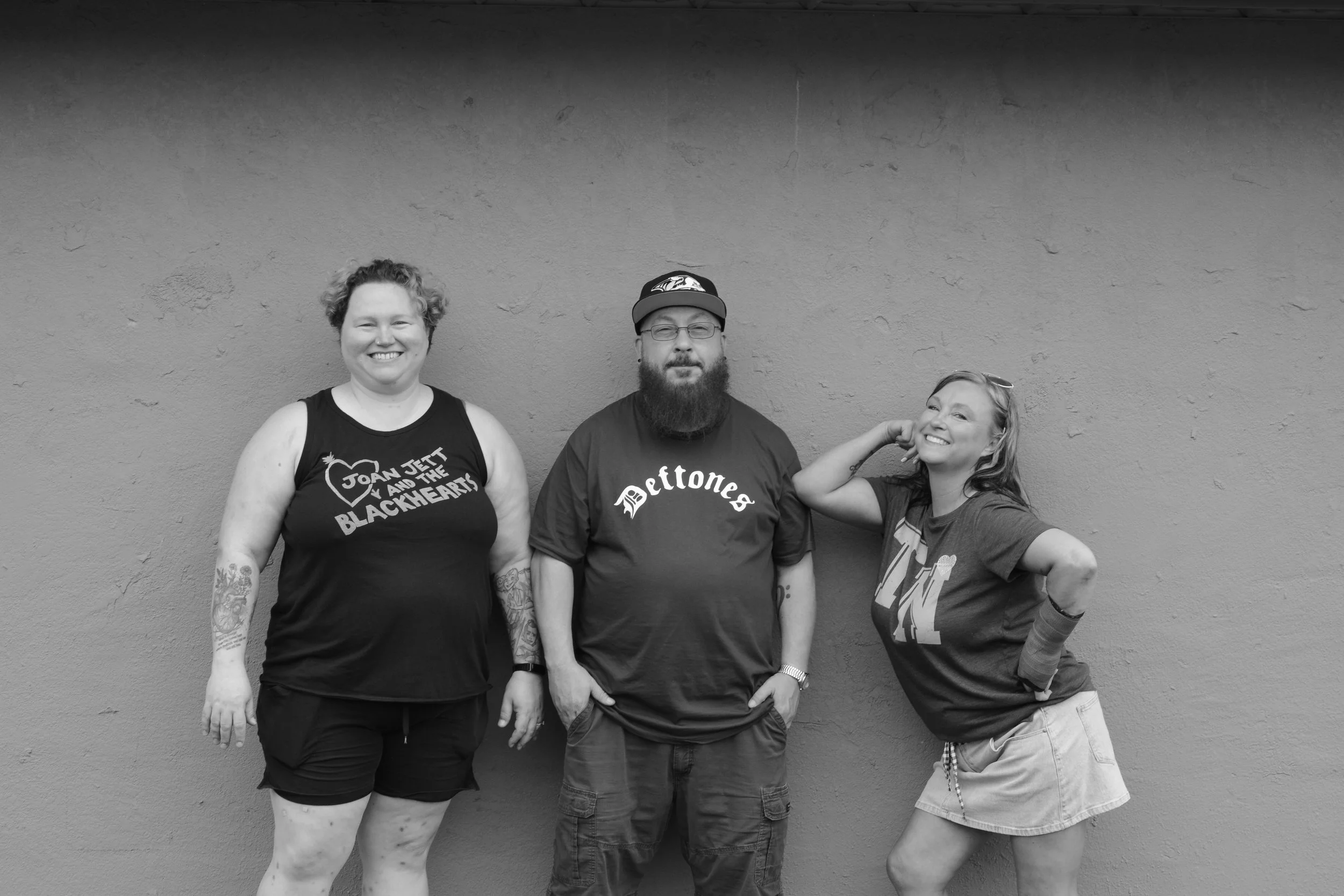 Three people standing in front of a plain wall, smiling and posing for the photo. The person on the left has short, curly hair, tattoos on their arms, and is wearing a sleeveless top. The person in the middle has a beard, glasses, and is wearing a ba