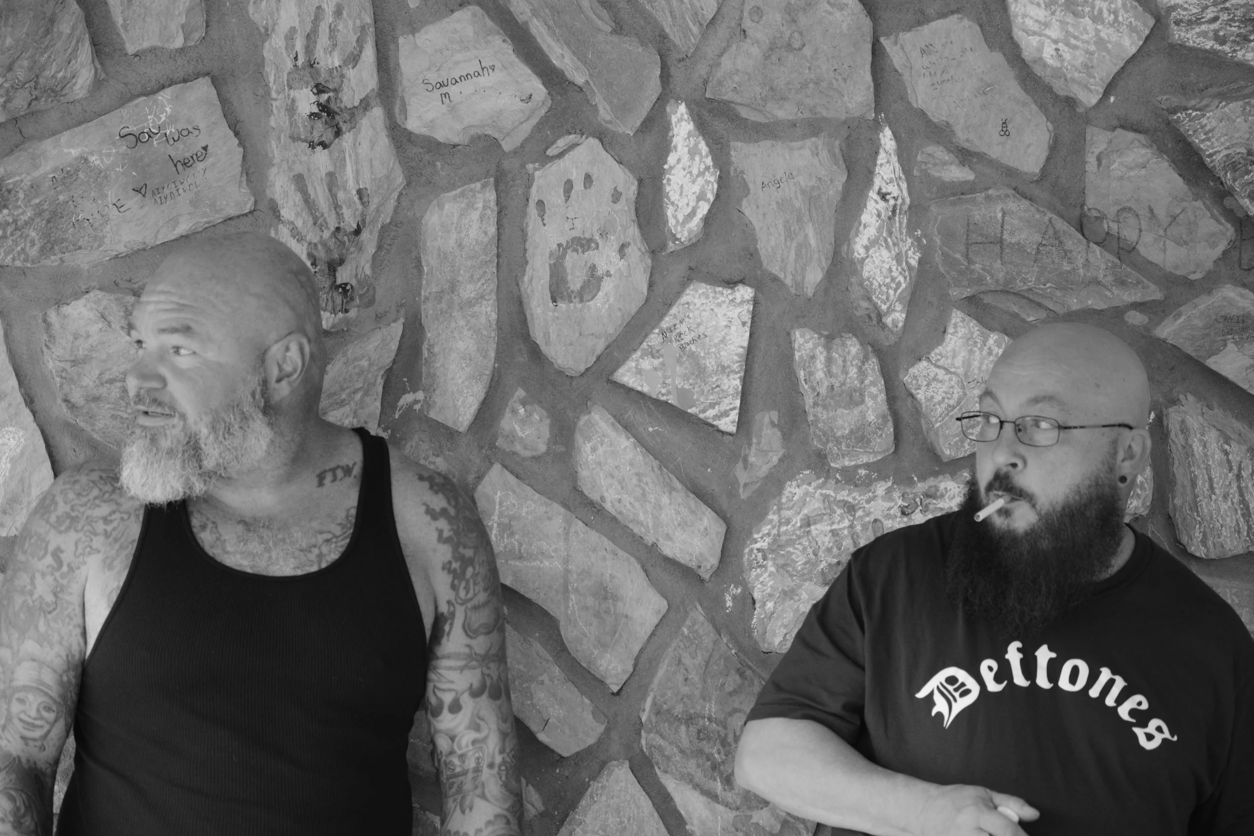 Two tattooed men with beards sitting against a stone wall, with one smoking and both looking away from each other.