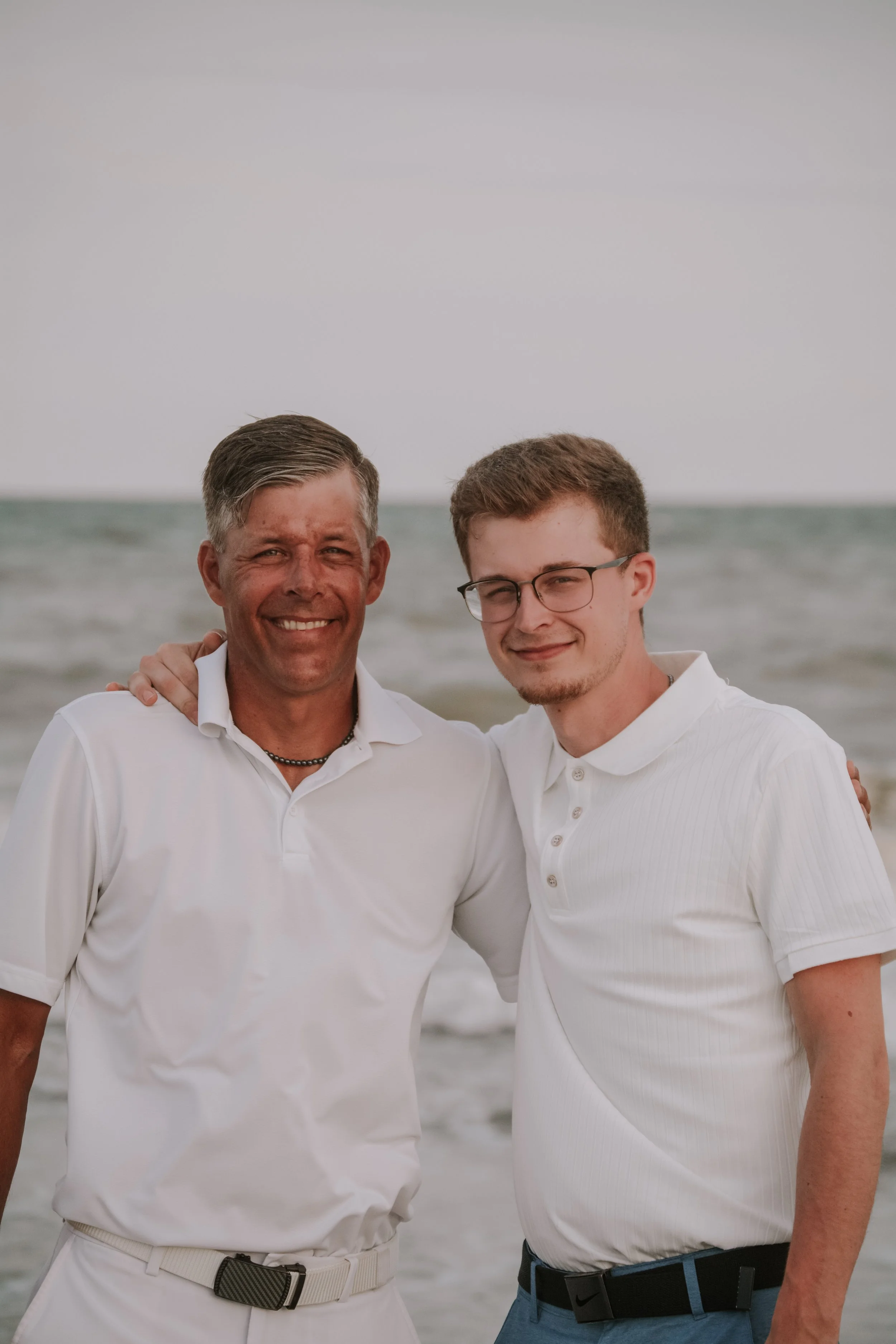 Two men standing on a beach, smiling. The man on the left has short gray hair, tan skin, and is wearing a white polo shirt. The man on the right has brown hair, glasses, and is wearing a white polo shirt. Overcast sky and ocean waves are in the backg