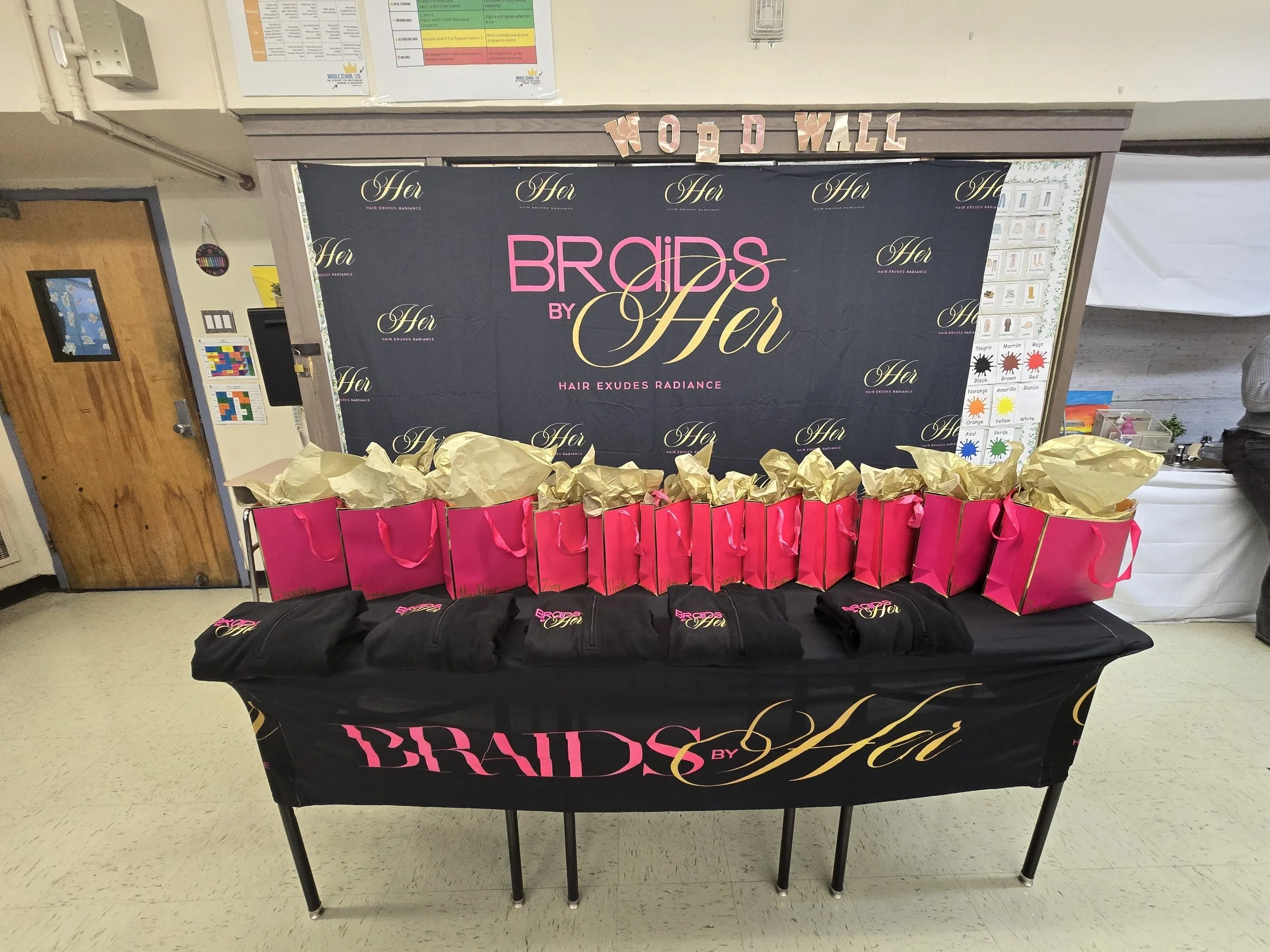 Table display with pink gift bags, brown paper wrapping, black items with pink and gold text, and a backdrop with logos and text about brides and hair.