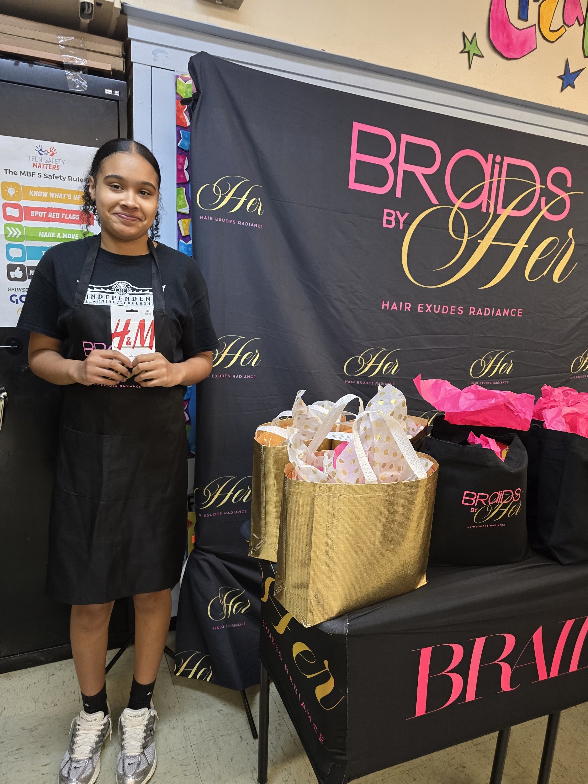 A young woman standing next to a table with gift bags, smiling, in front of a sign that says 'Braids by Her'. She is wearing a black t-shirt, a black apron, white sneakers, and black socks.
