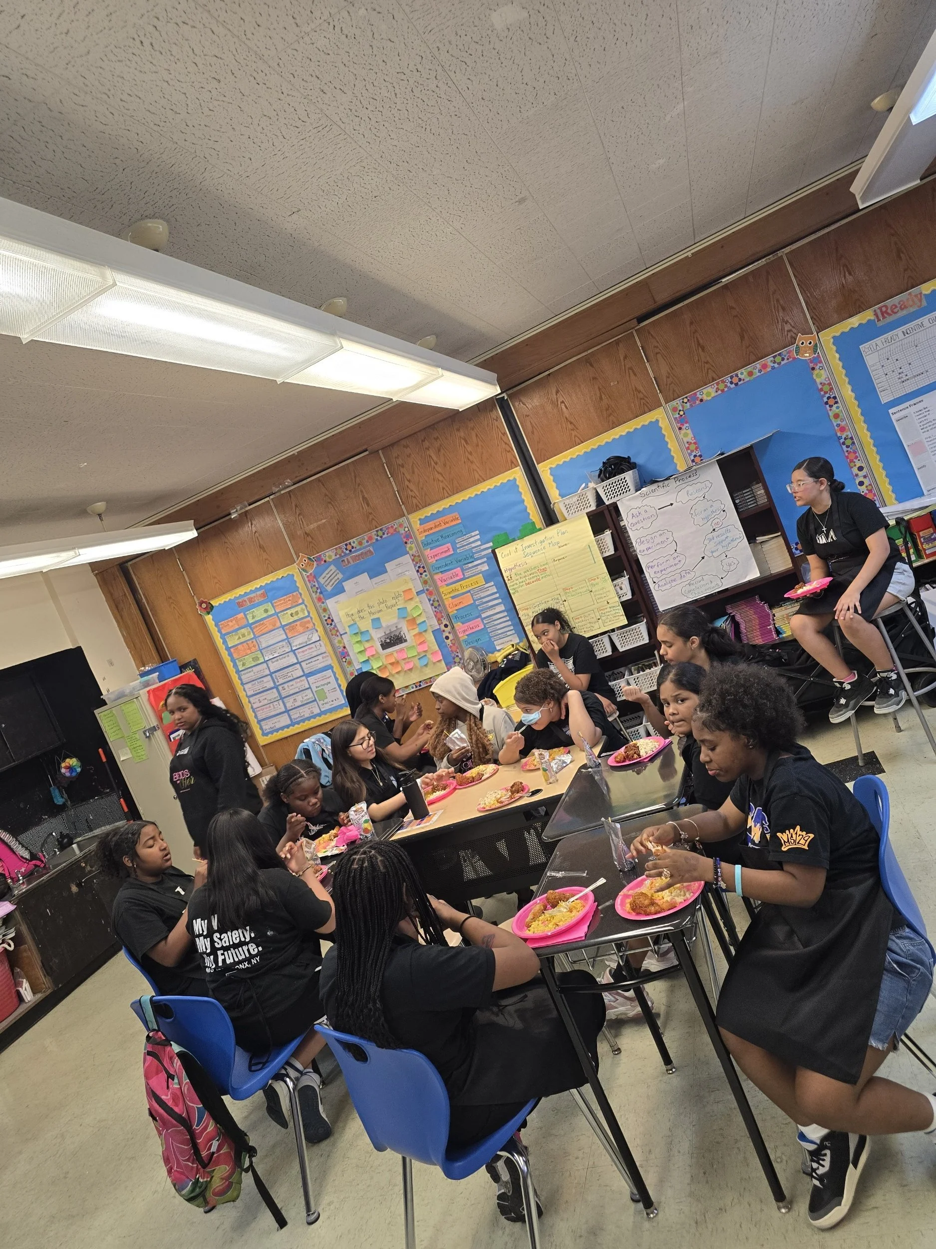 Students gathered in a classroom sitting at tables, eating lunch from pink plates, engaging in conversation. The classroom has educational posters and bulletin boards on the walls, and a woman standing on a chair is overseeing the students.