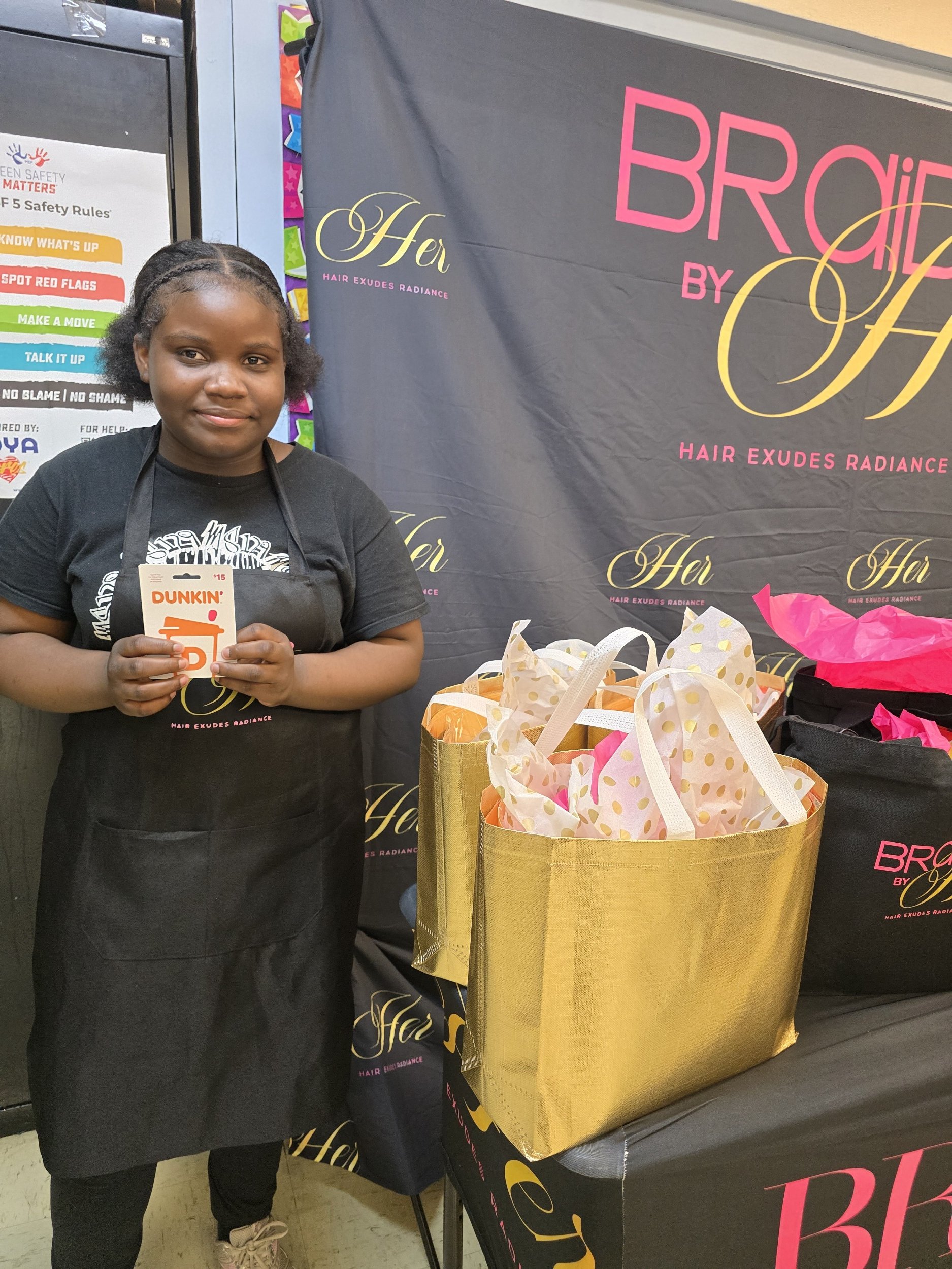 A young girl with braids, wearing a black apron over a black t-shirt, holding a Dunkin' gift card, standing next to a large gold gift bag filled with pink and white tissue paper. Behind her is a black display with pink and gold text reading "Her" and "Browt by Her," and a colorful hand safety poster.