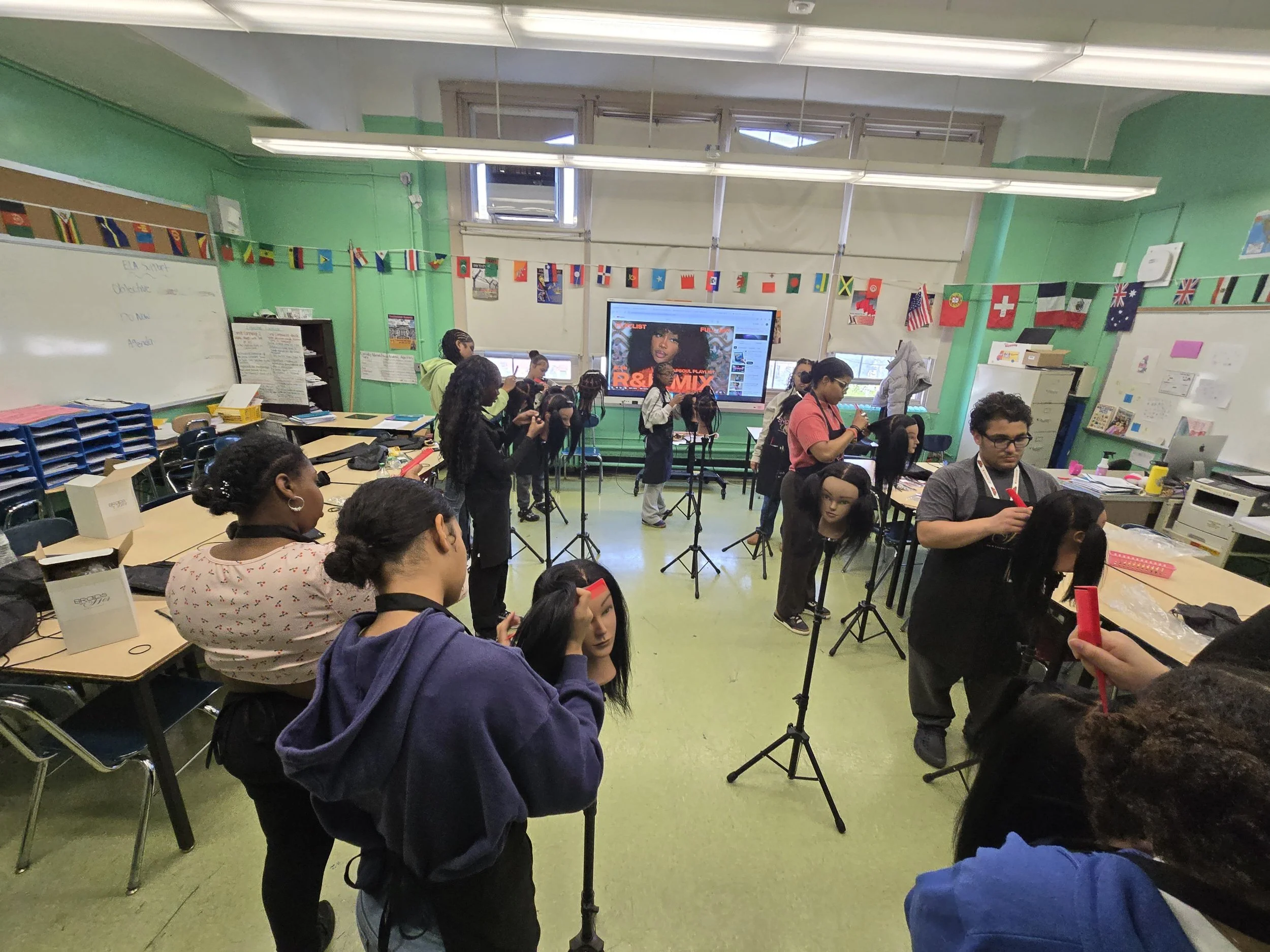A classroom filled with students, some standing and some sitting, while they use their phones or look around. The classroom has green walls, decorated with international flags and posters. There is a large screen at the front displaying a music playlist, with mannequins and musical equipment in the foreground.