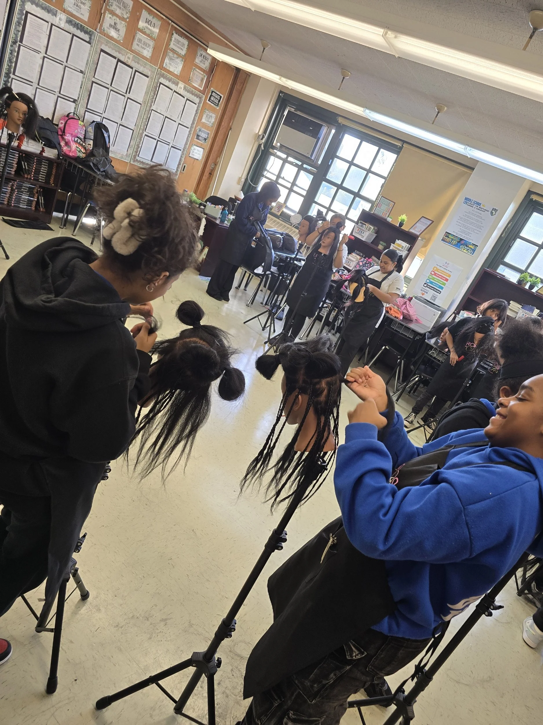 A classroom with students and hairstylists, some students are seated and being styled, with hairstylists working on hair, and backpacks and supplies on tables and shelves. There are posters and decorations on the walls and large windows letting in su