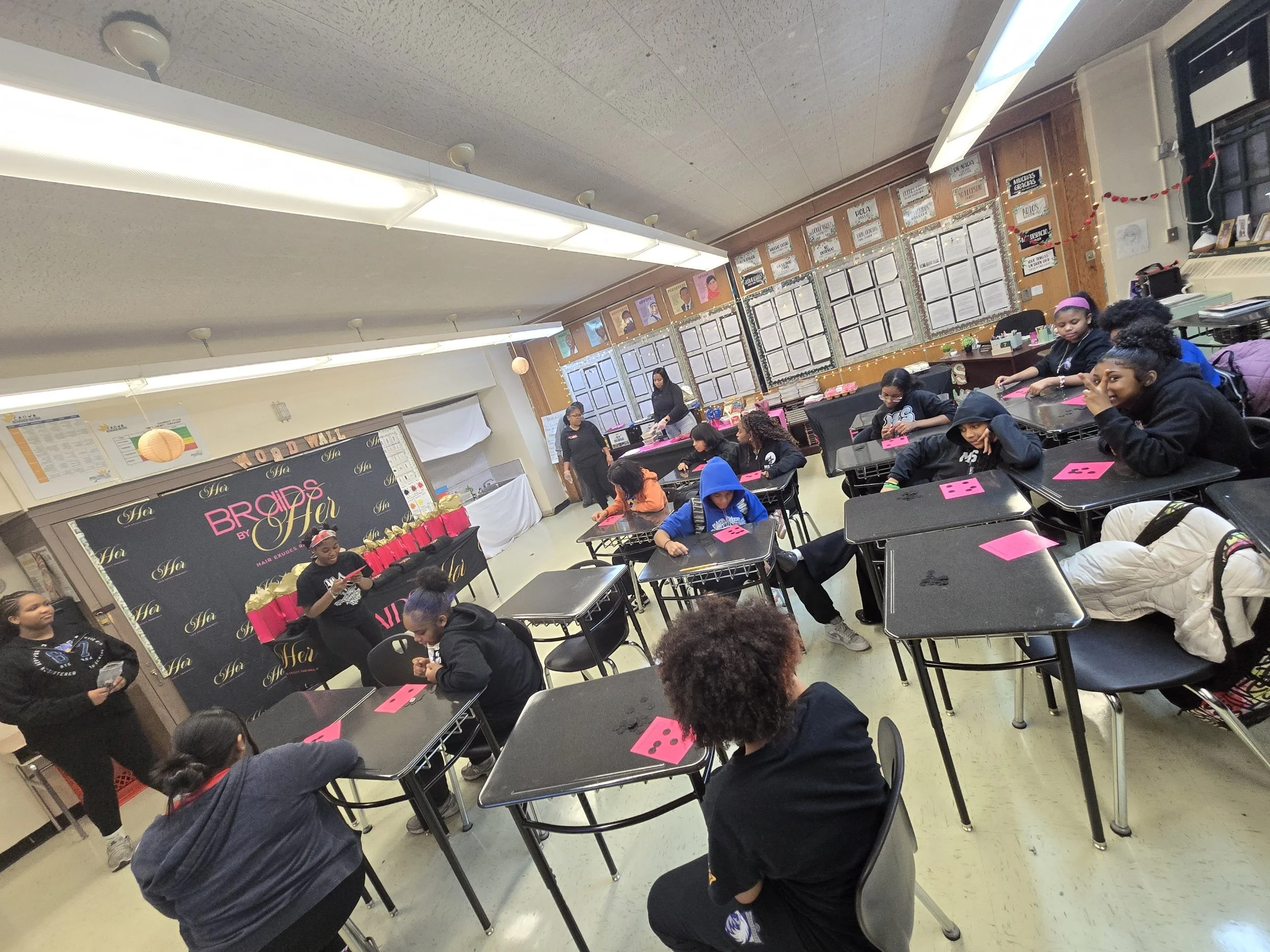 Students sitting at tables in a classroom, some posing and smiling, others focused on activities, with bulletin boards and decorations on the walls.