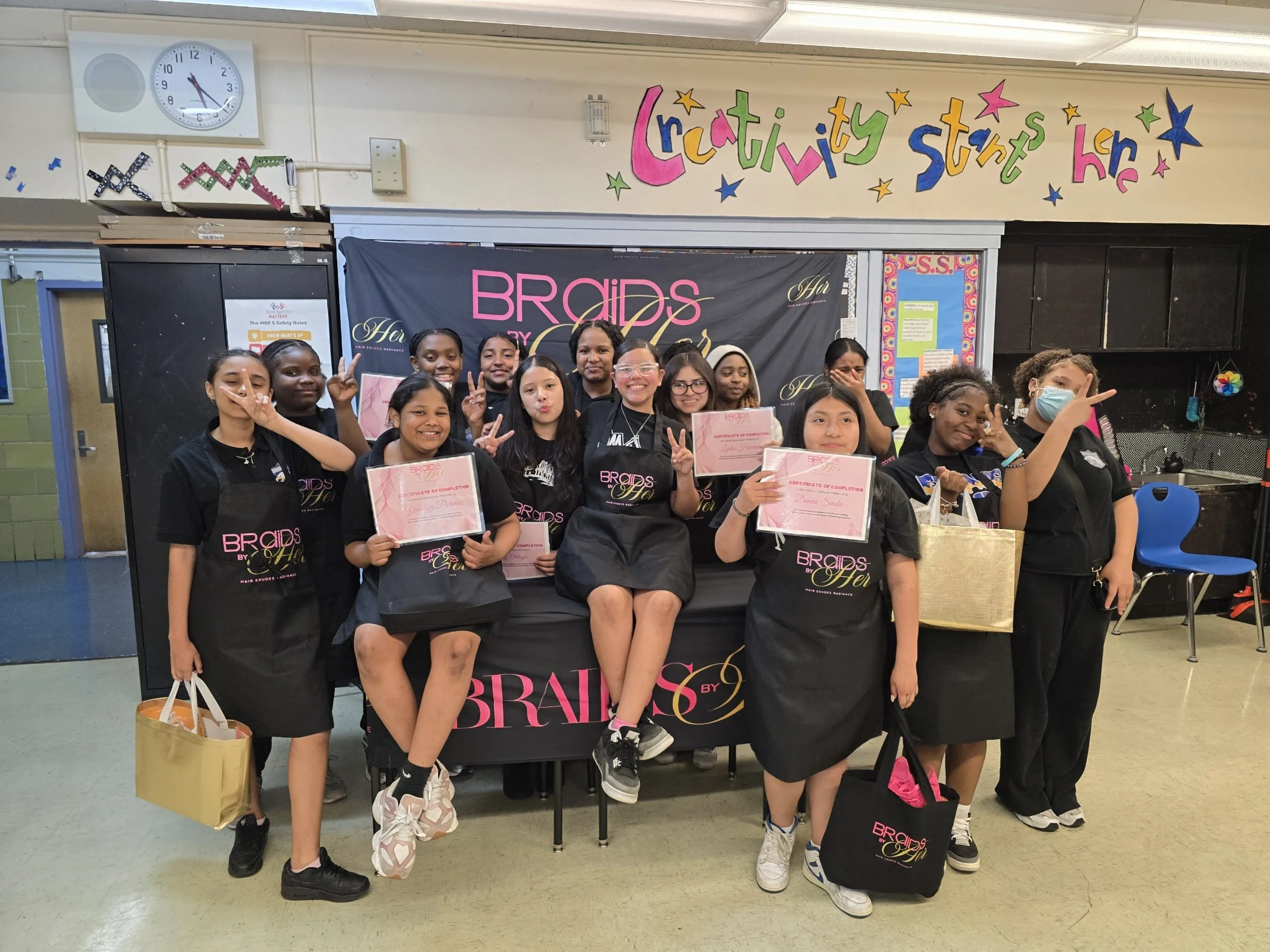 Group of young girls holding certificates and smiling at a celebration event in a school gymnasium. They are wearing black shirts with the word 'Braids' in pink and gold lettering. A large banner behind them reads 'Braids by Her' and there is a sign 