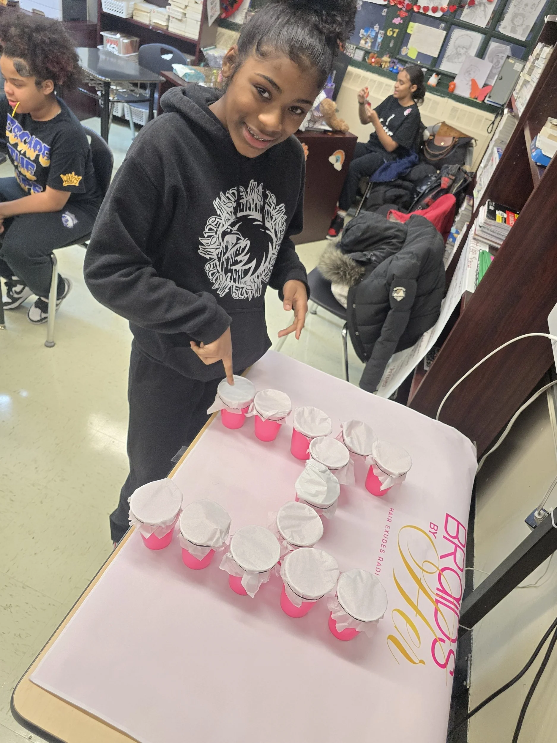 A young girl with braces smiling and pointing at a row of pink cups with white paper covers on a table, with other children and classroom decorations in the background.